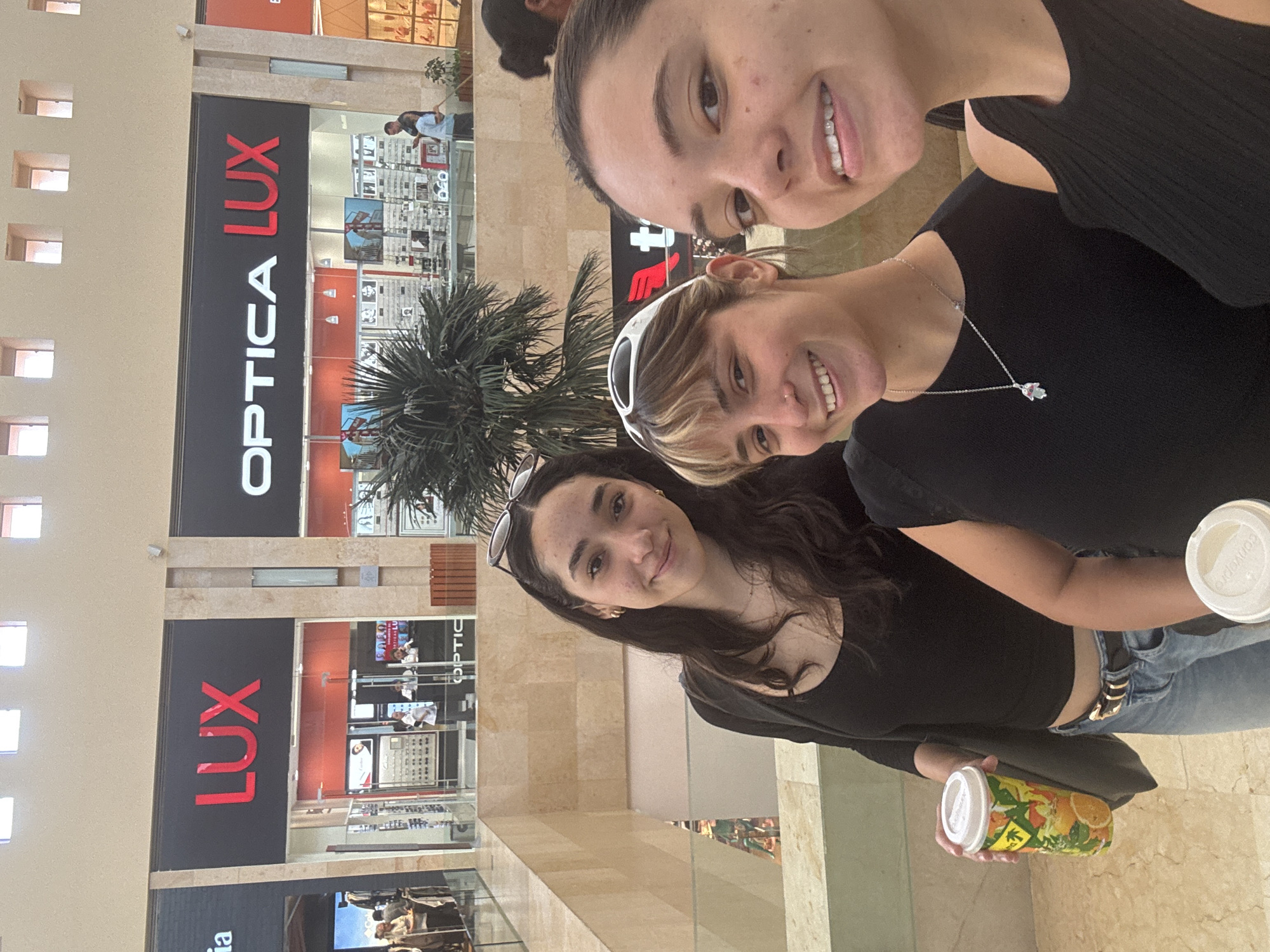 Three young women are posing for a selfie in what appears to be a shopping mall. The mall is located in Puebla, Mexico, as indicated by the "OPTICA LUX" sign, which is a business known to operate in Puebla, Mexico. The women are smiling at the camera. The woman in the front holds a container with an orange design. One woman has sunglasses on her head. The woman in the middle is wearing a silver necklace. "OPTICA LUX" is visible in the background. The mall has a multi-story design with tan-colored walls and modern architectural elements. There are people and merchandise visible inside the store behind the women.