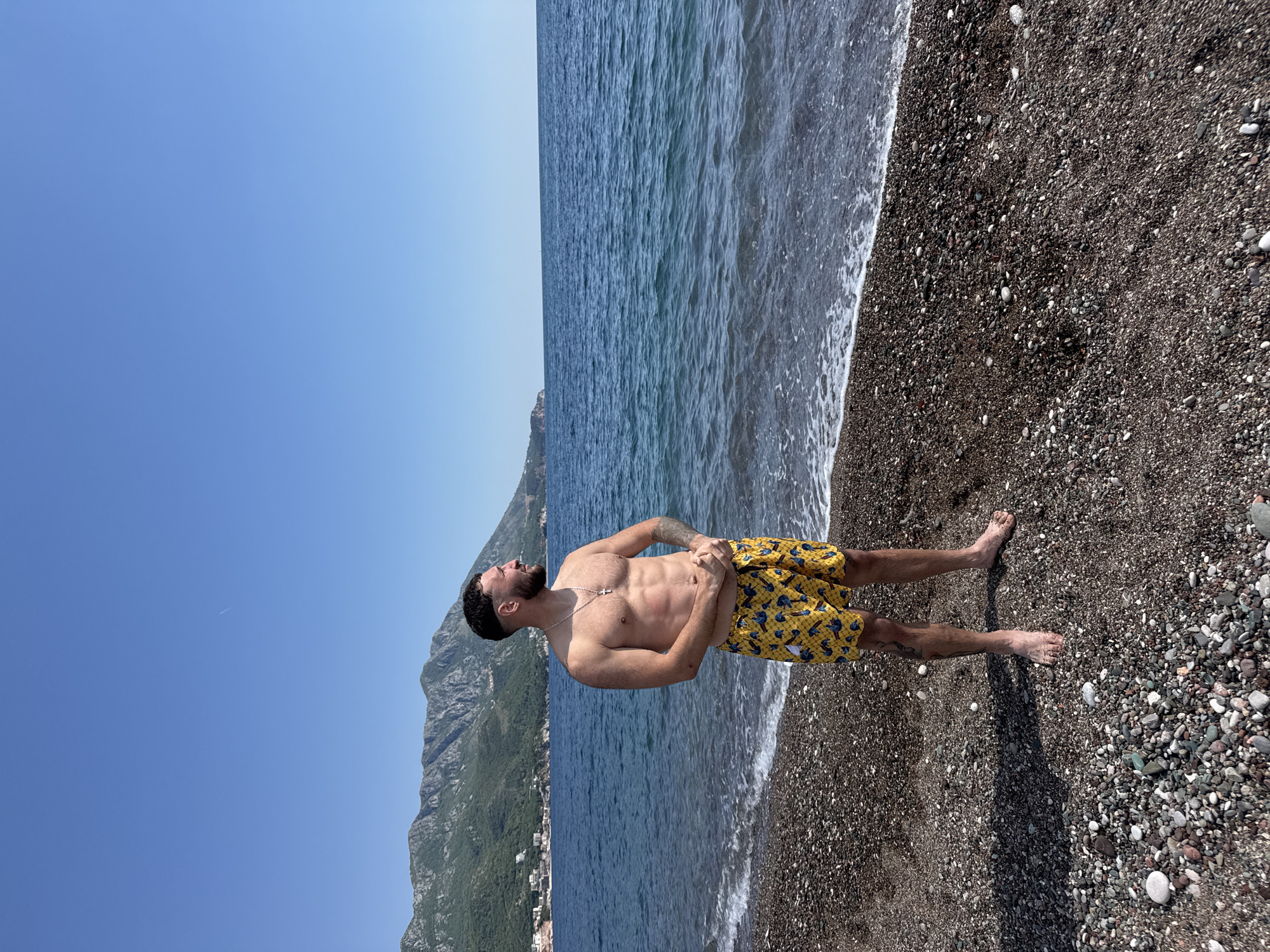 A medium shot of a man with a beard standing on a pebble beach, facing left. He is shirtless and wearing yellow swimming trunks. He has his hands on his abdomen and is looking upward. Behind him is the ocean with a mountain backdrop. The sky is a clear blue. The location is unknown.