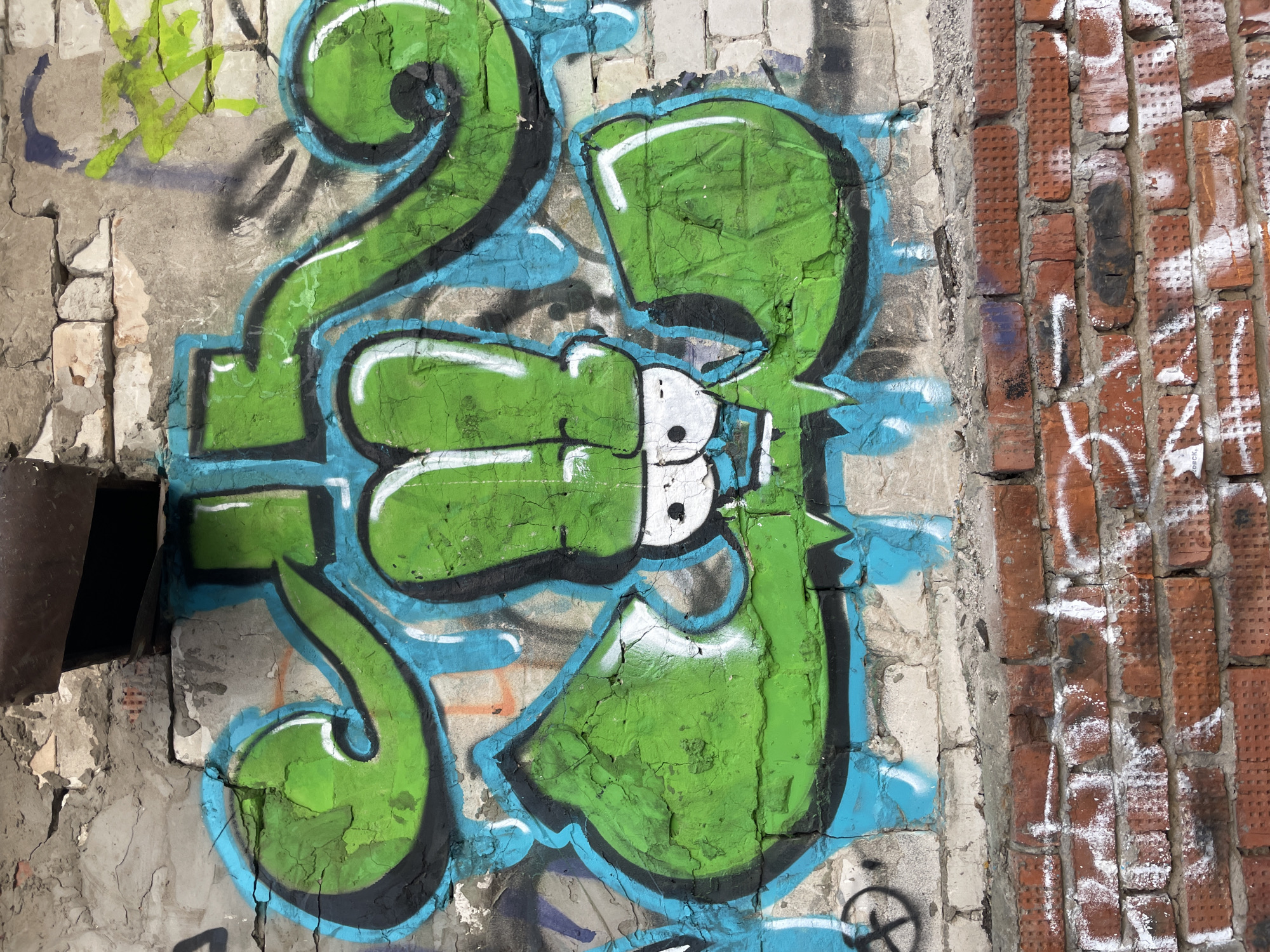 The image displays a vibrant graffiti artwork on a heavily weathered urban wall, situated in what appears to be Izhevsk, Russia. The wall is divided into two distinct sections: a crumbling concrete surface on the left and a red brick wall on the right.
The central feature is a large, stylized graffiti piece depicting the numbers "2" and "3". Rendered in bright green with bold black outlines and a light blue drop shadow, the artwork dominates the concrete section. The number "3" is particularly eye-catching, incorporating a white, cartoonish face or skull-like element with black eyes. The green paint shows signs of cracking and texture, indicating the age of the art or the wall's rough surface.
The concrete wall itself is severely dilapidated, with prominent cracks, peeling plaster, and sections where white bricks are exposed beneath. A dark metal pipe or conduit is partially visible on the far left edge. Faint, older graffiti in lighter green and purplish-blue tones, along with a circular symbol (possibly a peace sign), can be seen underneath or alongside the main piece.
The adjacent brick wall on the right is composed of old, textured reddish-brown bricks. This section is also marked with various white and black spray-painted tags and illegible scribbles. Among these, the word "KOCK" is discernible in white spray paint.
The scene is brightly illuminated, suggesting daytime conditions that highlight the intricate textures of the decaying walls and the vivid colors of the street art. The overall impression is one of urban decay infused with expressive artistic intervention.