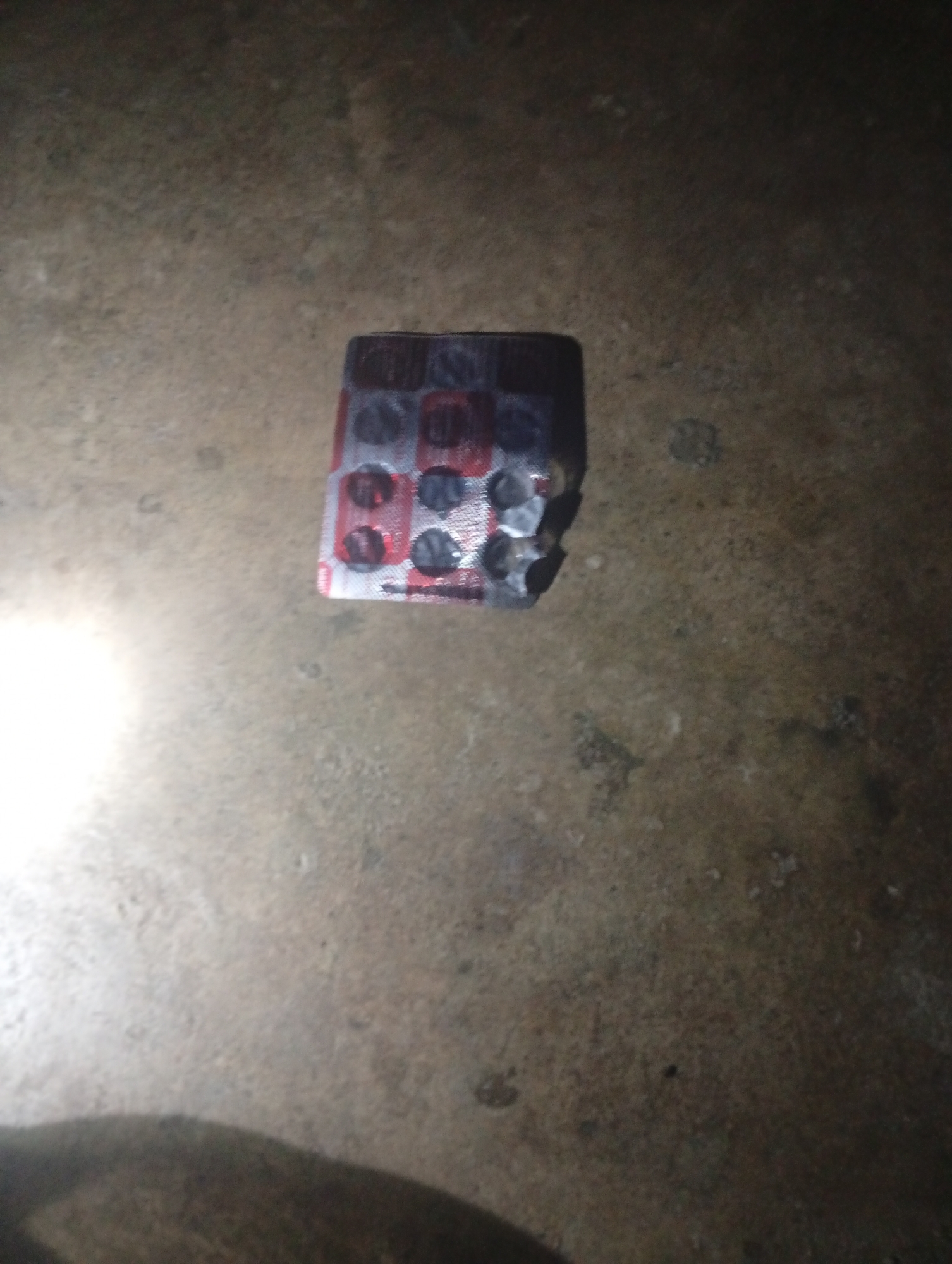 A partially used blister pack of red and white pills lies on a dark, rough, textured surface that appears to be concrete or asphalt. The blister pack is oriented horizontally and is slightly damaged on the right side. The surface is uneven with small pebbles and imperfections visible. The lighting in the scene is dim, casting a shadow to the lower left of the blister pack. The location is described as Unknown, Unknown.
