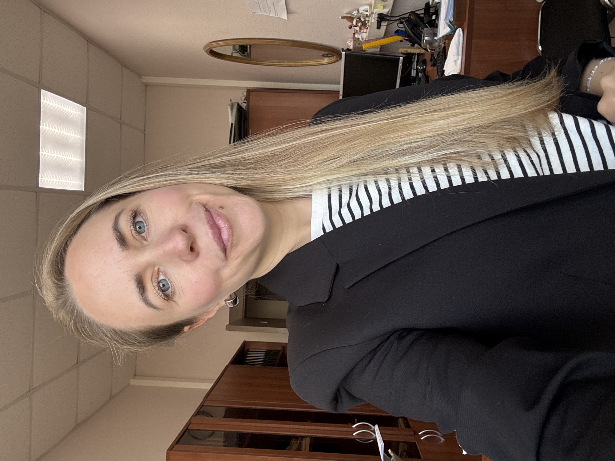 The image displays a woman taking a close-up portrait, likely a selfie, within what appears to be an office or workplace in Chaikovsky, Russia. The woman has long, straight blonde hair, which is swept over her left shoulder. She has light blue eyes and a subtle, friendly smile, looking directly into the camera. She is dressed in a black blazer over a white and black horizontally striped top. A silver bracelet adorns her right wrist, and she wears small hoop earrings.
The background reveals a functional office environment. The ceiling features acoustic tiles and a prominent, illuminated rectangular light fixture. The walls are a light, neutral color. Behind the woman, a dark wooden cabinet or shelving unit is visible. On a desk or table surface, various office items are scattered, including a black computer monitor, a landline phone, papers, a clear glass, and a white cloth, suggesting a lived-in work area. A small, oval-shaped mirror with a gold frame hangs on the wall. The scene is brightly lit by the artificial ceiling light, and no specific time of day is discernible beyond being indoors. No discernible text is visible in the image.