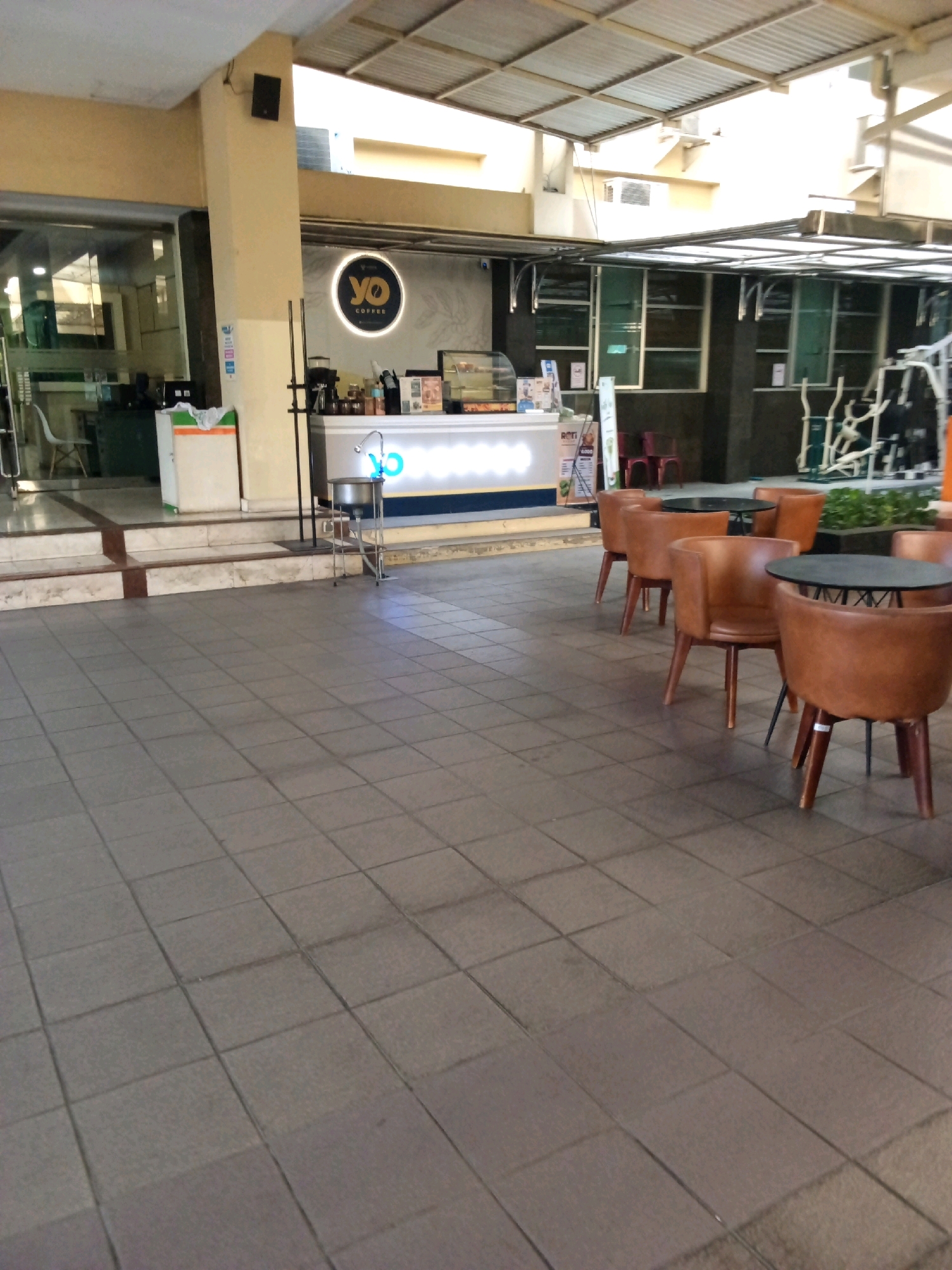 An outdoor seating area for a coffee shop is depicted. The coffee shop, named "YO COFFEE," has a counter with coffee-making equipment and displays of food items. Adjacent to the coffee shop are several brown leather chairs and round black tables. In the background, exercise equipment is visible, suggesting the coffee shop might be located in a gym or fitness center. The ground is paved with gray square tiles. The scene appears to be in a commercial area.