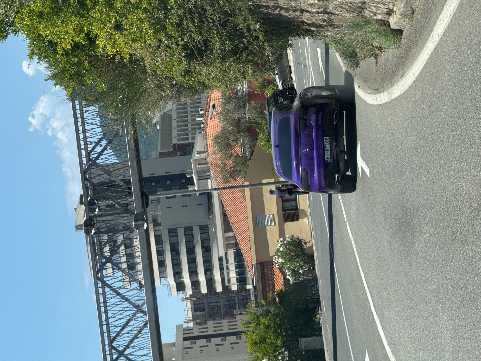 A purple Mercedes-Benz sedan is traveling on a paved road in Rafailovići, Montenegro. The car, bearing the license plate "BD BT 168," is seen from behind as it curves to the right. A person wearing a dark shirt and a cap stands on the side of the road near a yellow building with orange-tiled roofs. In the background, modern buildings and an overpass structure are visible against a clear blue sky. Lush green vegetation, including trees and bushes, lines the right side of the road, with a stone wall bordering the vegetation. The road has white lane markings and directional arrows.