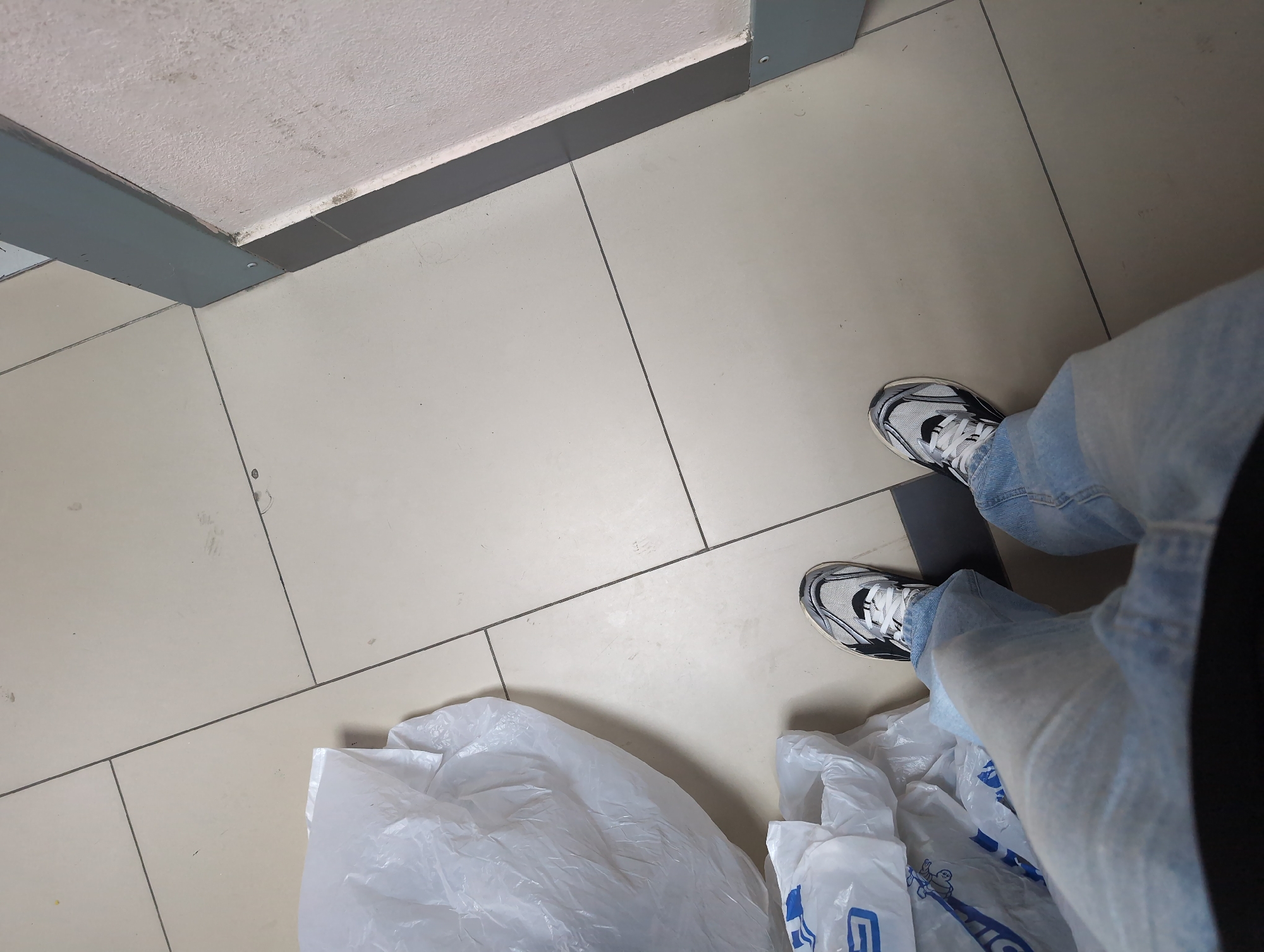 The image shows a pair of feet wearing light-wash jeans and athletic shoes, standing on a tiled floor. The person is positioned near several white plastic shopping bags. The background features a wall with a contrasting texture and a gray trim. The lighting appears to be artificial, casting subtle shadows on the floor. There is no visible indication of the location being in Krasnodar, Russia.