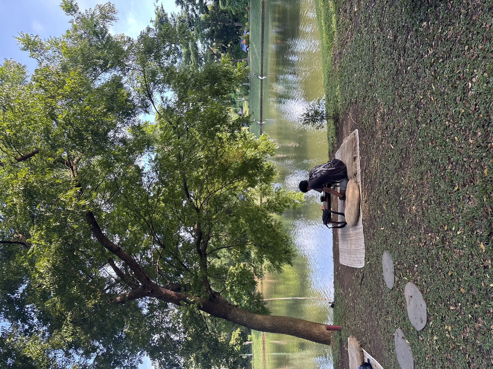 A person is setting up a pour-over coffee station next to a tree on the shore of a pond. The scene takes place in Pecatu, Indonesia, indicated by the tropical environment. The person wears a dark, patterned shirt and kneels on a woven mat. A dark backpack rests on a stool next to the coffee apparatus. The pond's surface reflects the sky and surrounding trees. Stepping stones are on the grass near the mat.
