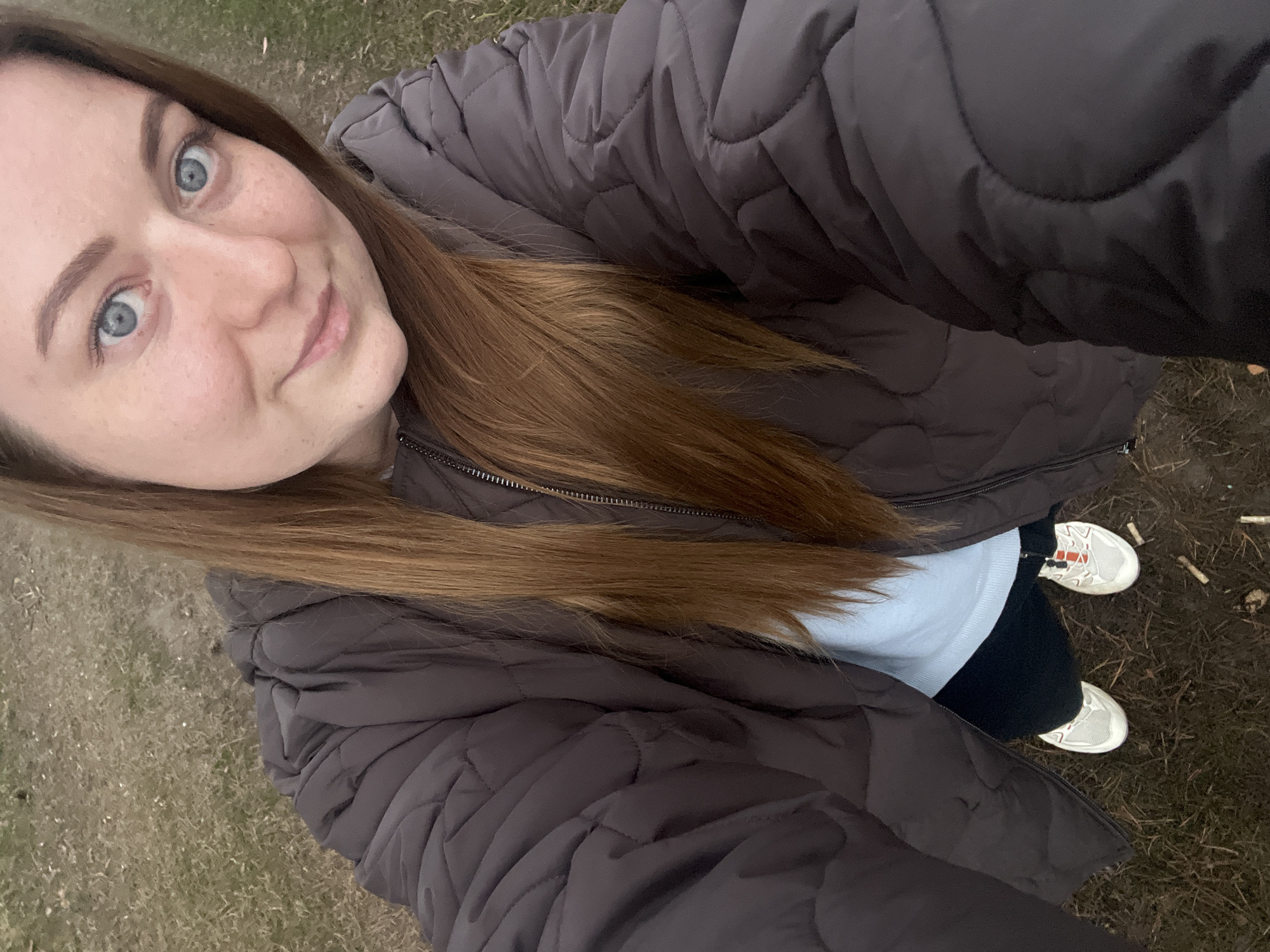 A selfie of a young woman, possibly in her 20s, taken from a high angle looking down at herself, with her face occupying the upper left of the frame. She has fair skin, light blue eyes, and long, light brown hair. She is wearing a brown quilted jacket over a light blue sweater and dark leggings. Her feet are visible at the bottom right, shod in white sneakers. The ground underfoot appears to be covered in sparse, dry grass. This photo was taken outdoors, seemingly in the city of Razdolye, Russia.