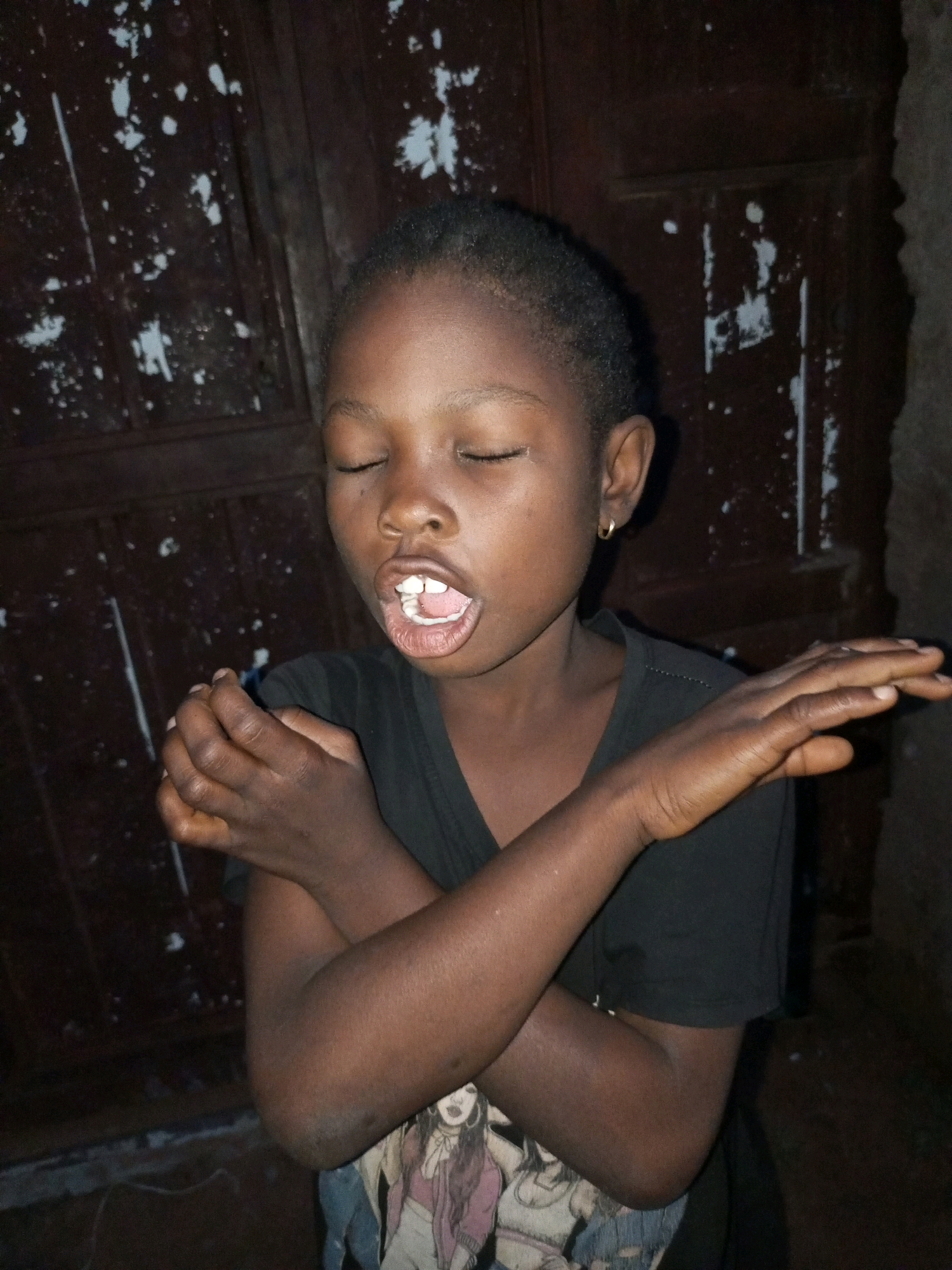 A young Black girl with closed eyes and an open mouth, showing her teeth, is pictured. She is wearing a black t-shirt and a gold hoop earring. Her arms are crossed in front of her. She is standing in front of a dark brown, weathered wooden door with white paint splotches. The environment appears to be outdoors, with some rubble and a darker, rough wall visible to the right. The lighting suggests it is nighttime or a dimly lit interior space.