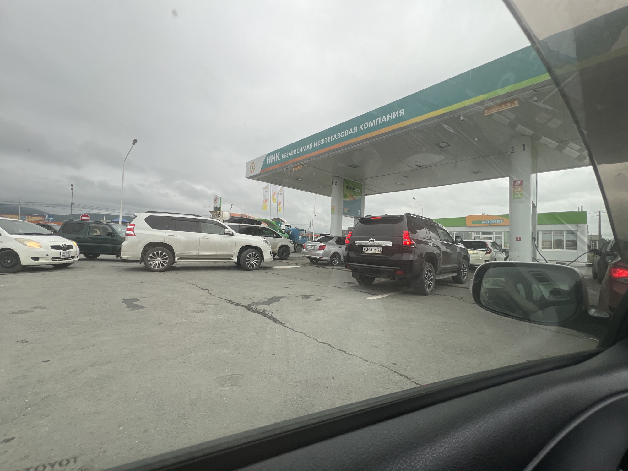 A queue of vehicles is present at a gas station. A white Toyota Land Cruiser is in the foreground, followed by a dark SUV, a silver car, and a white SUV. The gas station canopy is green with yellow trim, and bears the logo "ННК НЕФТЕГАЗОВАЯ КОМПАНИЯ". The sky is overcast. The scene appears to be in Yuzhno-Sakhalinsk, Russia, based on the license plates and gas station signage.