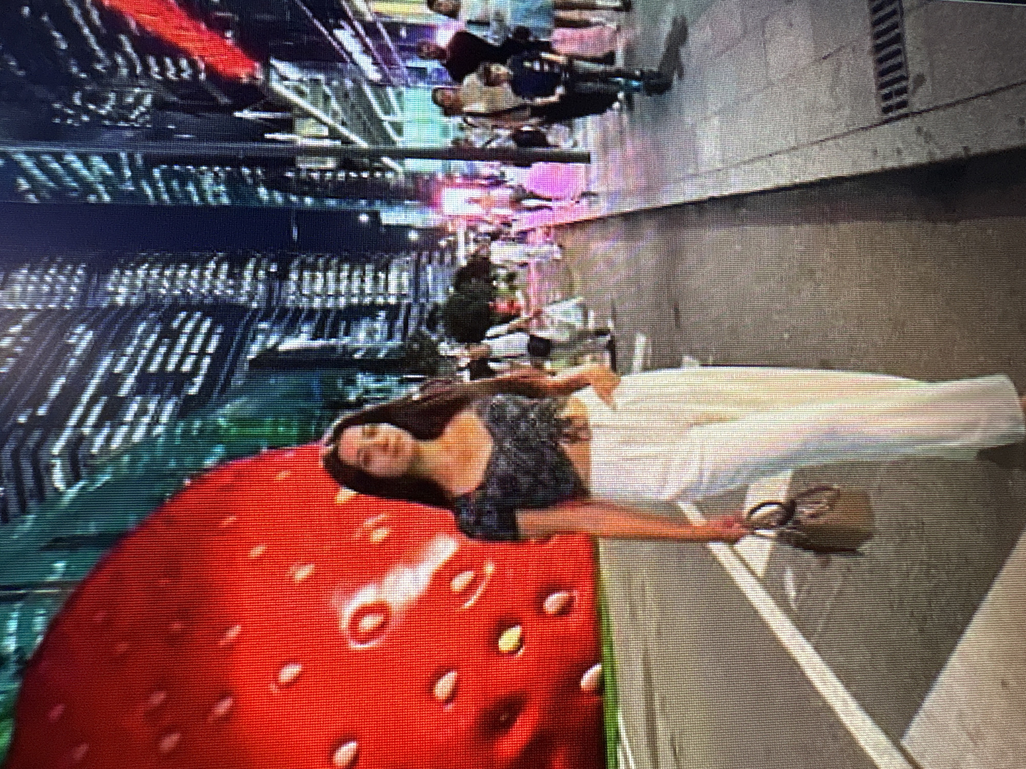A woman is walking on a city street at night, in front of a large illuminated strawberry sculpture. She is wearing a dark patterned crop top and white pants, carrying a small brown handbag. The background shows illuminated skyscrapers and other people walking on the sidewalk. The scene suggests a lively urban environment with a festive or art installation. The image quality is low, with visible pixels, making precise identification of individuals or specific architectural details difficult. The location is not identifiable as Moscow, Russia, based on the visual information provided.