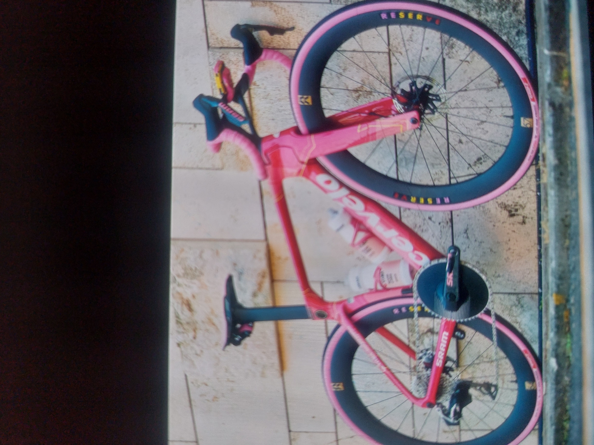 A pink Cervelo bicycle with black tires and pink sidewalls, bearing the word "RESERVE" on the sidewalls, is laying on a tiled surface next to a metal railing. The bicycle has SRAM components and is equipped with a water bottle. The location is unknown; there is no information about Tsarevo, Bulgaria visible in the image.