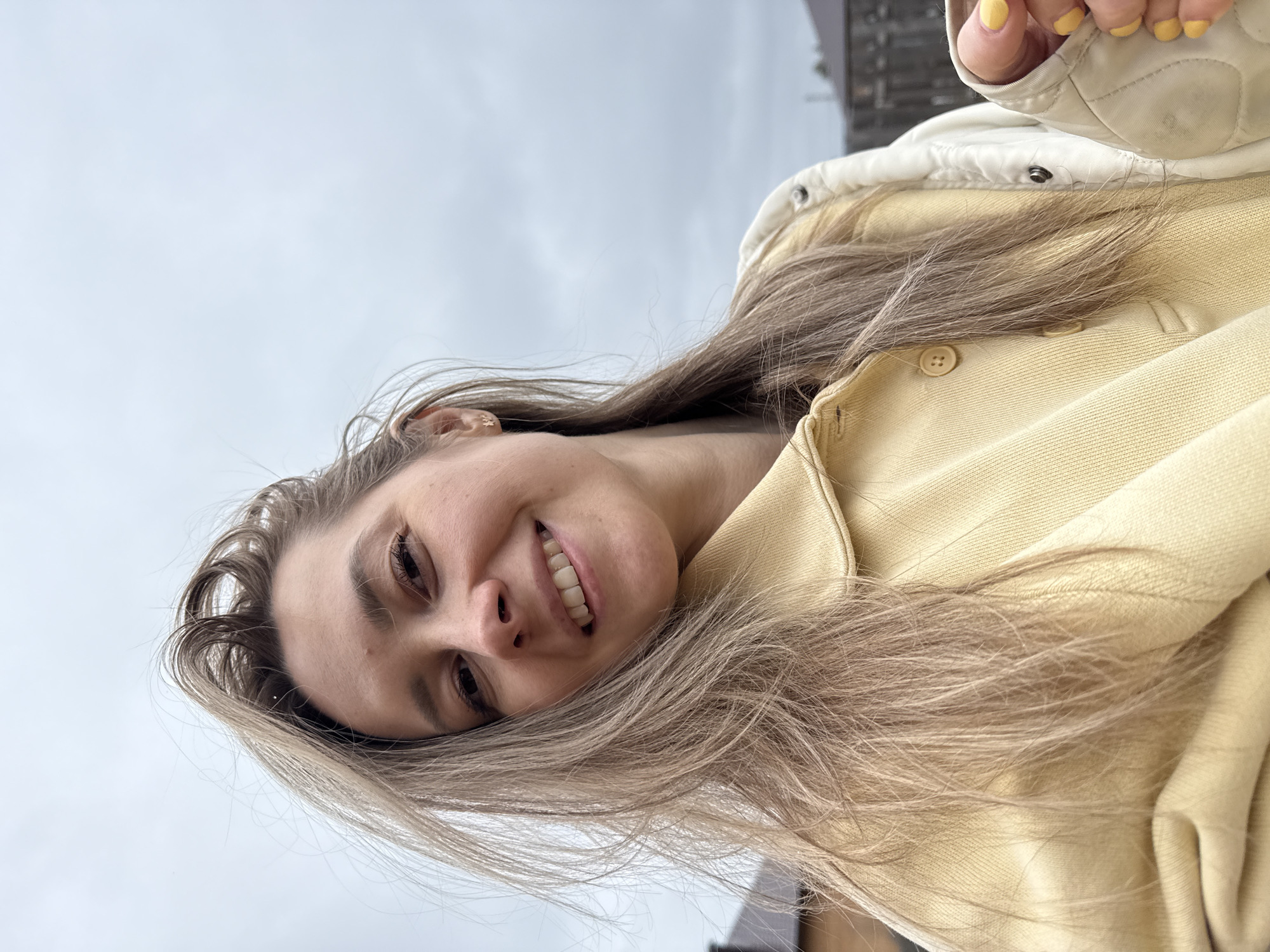 A young woman is featured in a close-up, sideways selfie taken outdoors in Chaikovsky, Russia. She is smiling broadly at the camera, her mouth open slightly to reveal her teeth. Her long, light brown or blonde hair cascades around her face and shoulders. She is dressed in a light yellow jacket or hoodie, layered with an off-white quilted vest. Her fingernails are painted a vibrant yellow, visible near the top right of the frame where her hand is positioned.
The image is captured from a low angle, looking upward, with the woman's face and upper body filling the majority of the frame. The background consists of a bright, uniformly gray, overcast sky, indicating cloudy weather. Some indistinct, dark structures, possibly buildings, are faintly visible in the far background on the right side. The woman appears cheerful and content. No text is visible in the image.