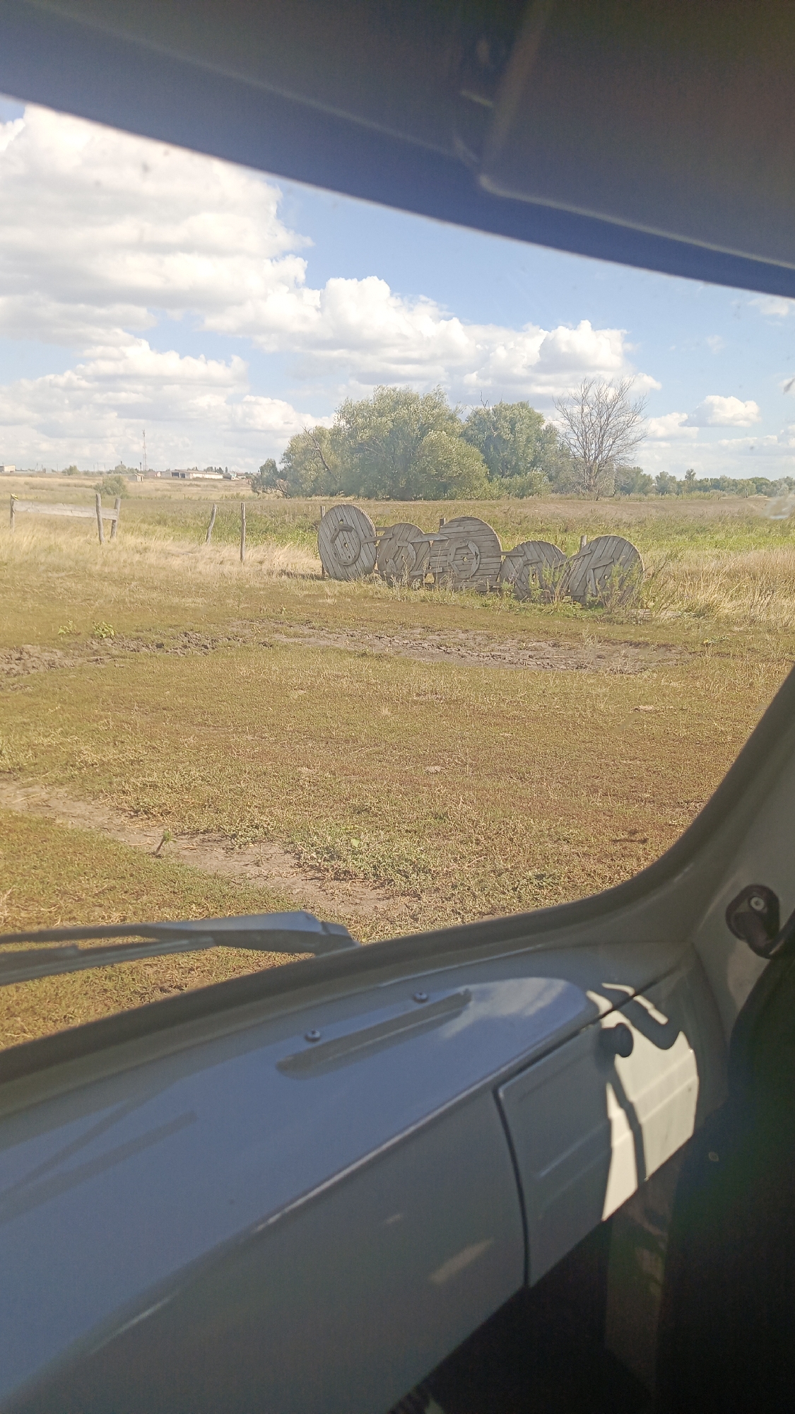 This is an outdoor, medium-distance shot taken from inside a vehicle, looking out through the windshield. The foreground shows a portion of the vehicle's interior, including the dashboard and windshield wipers. The background depicts a rural landscape under a partly cloudy sky. A field of dry grass occupies the middle ground, with a row of large, wooden spools, likely for cables or wire, positioned towards the right side. A dirt track or path runs through the field. In the distance, there are trees, a few buildings, and a transmission tower. The scene appears to be in a non-urban setting, possibly the village of Karyakino, Russia, given the context provided. No people are visible in the image.
