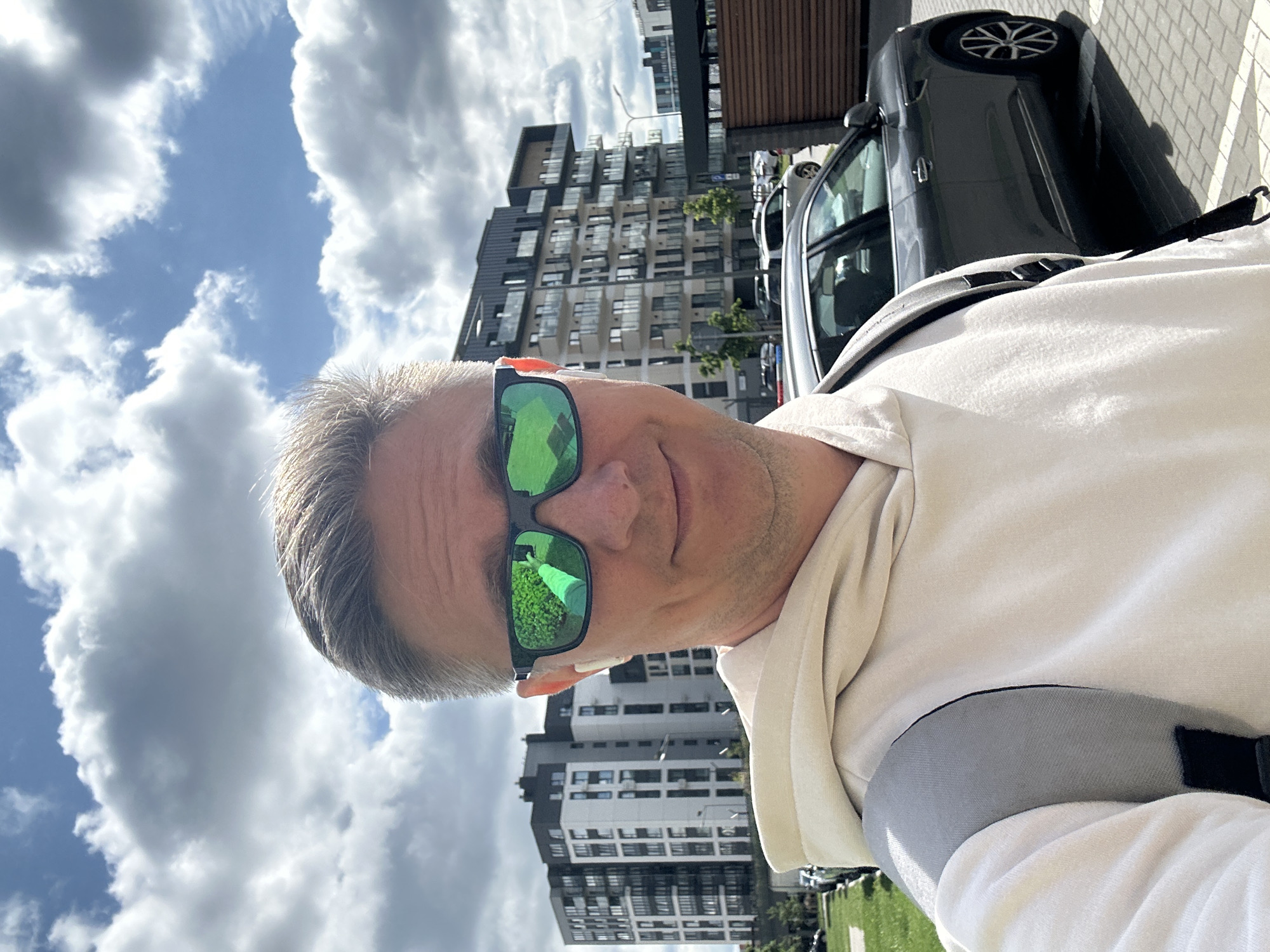 The image depicts a selfie of an adult male in an outdoor urban or suburban environment in Бараўлянскі сельскі Савет, Belarus. The man, with light-colored hair, is wearing green mirrored sunglasses and a white wireless earbud in his right ear. He is dressed in a light-colored (beige or cream) hooded sweatshirt, with a gray backpack strap visible over his right shoulder. A slight smile is present on his face. The reflection in his right sunglass lens shows a hand holding a smartphone, indicating the selfie-taking action. The background features modern multi-story residential buildings with white and dark gray facades, some with visible balconies, positioned to both the left and right of the subject. A dark gray SUV is parked on a paved surface to the right, with other vehicles partially visible further back. Green grass borders a paved area on the left. The sky is partly cloudy with patches of blue sky and numerous white and gray cumulus clouds.