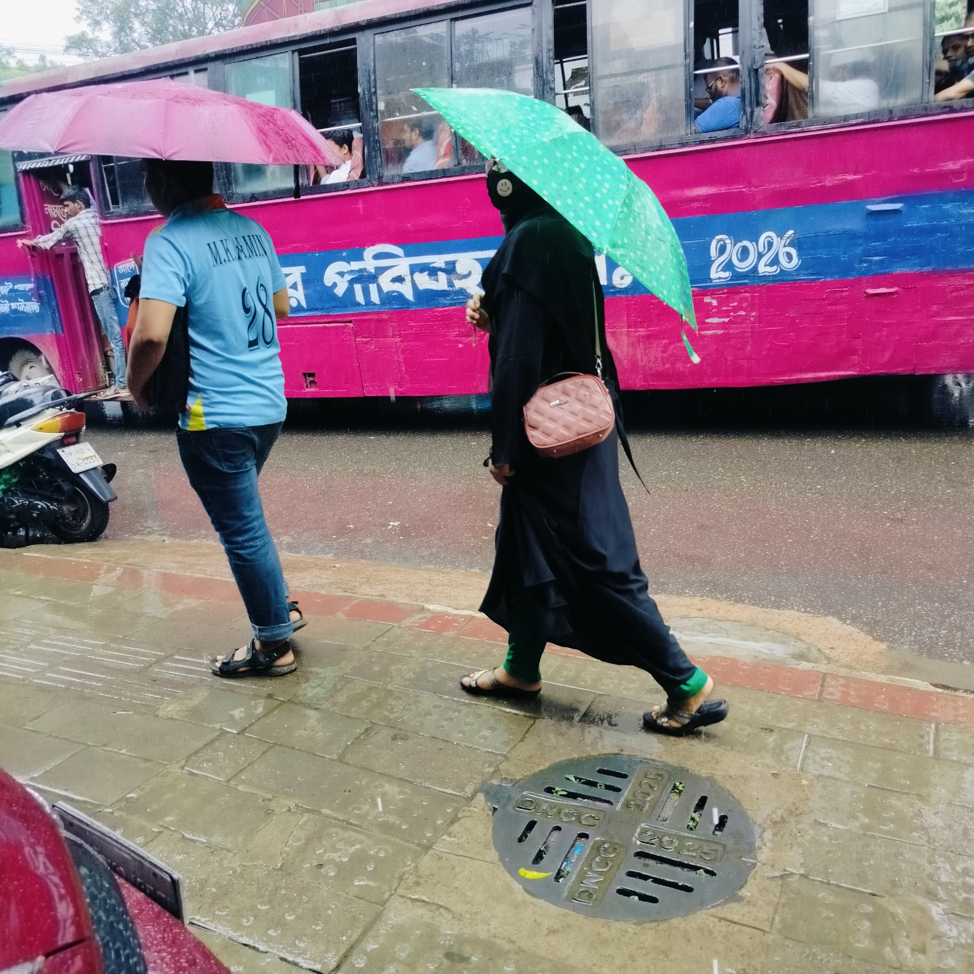 In Dhaka, Bangladesh, during rainfall, two individuals walk along a wet, tiled sidewalk adjacent to a pink and blue bus. The person on the left, wearing a blue t-shirt with "M.K.AMIN 28" on the back and blue jeans, walks under a pink umbrella. To their right, a person in a black abaya and green pants walks under a green, polka-dotted umbrella, carrying a small, quilted, pink handbag. Behind them, the bus displays the number "2026". A parked motorcycle is visible on the left side of the frame. A circular manhole cover with the inscription "DNCC 2025" is present on the sidewalk. The overall scene depicts an urban street during inclement weather.