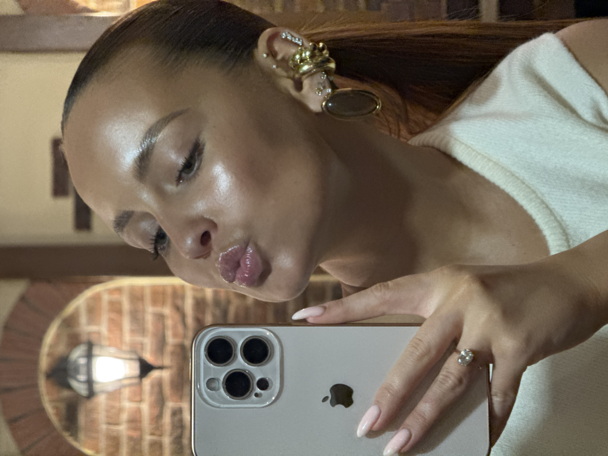 In a selfie, a young woman with light brown hair pulled back into a ponytail and wearing a white knit top is pouting her lips. She is holding a silver iPhone in her left hand, with her manicured pink nails visible. A diamond ring adorns her left ring finger. She has multiple piercings on her right ear, adorned with large gold earrings and smaller diamond studs. The background shows a brick wall with an arched opening, within which a vintage-style outdoor lantern is illuminated. The overall setting appears to be indoors with a brick wall and an arched doorway.