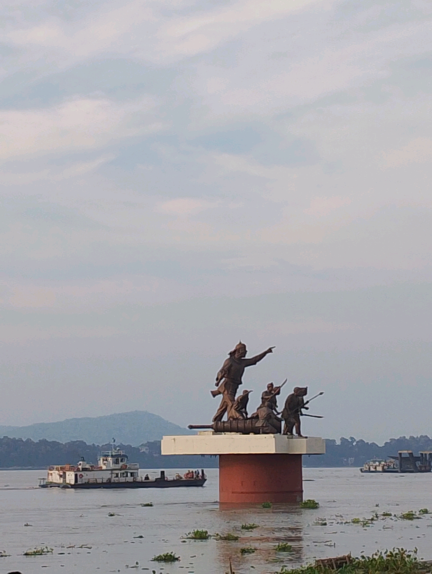 The image depicts a statue of warriors in a boat on a pedestal in the foreground, situated on a body of water, likely the Brahmaputra River, in Guwahati, India. The statue is bronze and features several figures, one prominently pointing forward. Behind the statue, a white ferryboat is visible navigating the water, with passengers on deck. In the distance, rolling green hills form the backdrop, and the sky is overcast with scattered clouds. The water surface is calm, with aquatic plants dotting its expanse. Another barge or boat can be seen in the far right of the image. The overall scene suggests a historical or cultural monument on the waterfront of Guwahati.
