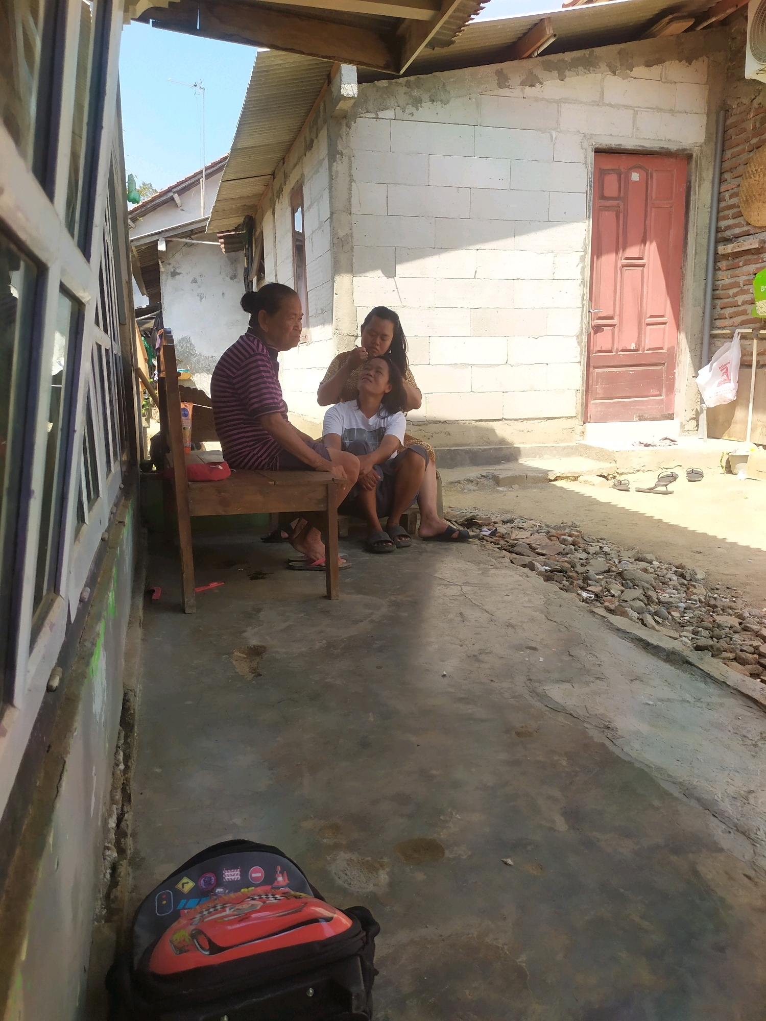 In Desa Penggalang Kecamatan Adipala, Indonesia, a woman in a striped shirt sits on a wooden chair while another woman, holding a child on her lap, rests her hands on the child's head. The child wears a white t-shirt. The scene unfolds outdoors, with a concrete walkway leading to a building constructed of white cinder blocks and a red door. To the left, a window with multiple panes is visible. The ground is a mix of concrete and dirt with scattered rocks. A child's backpack featuring a red car design rests on the ground near the bottom left of the frame. The environment appears to be a residential area with a clear sky.
