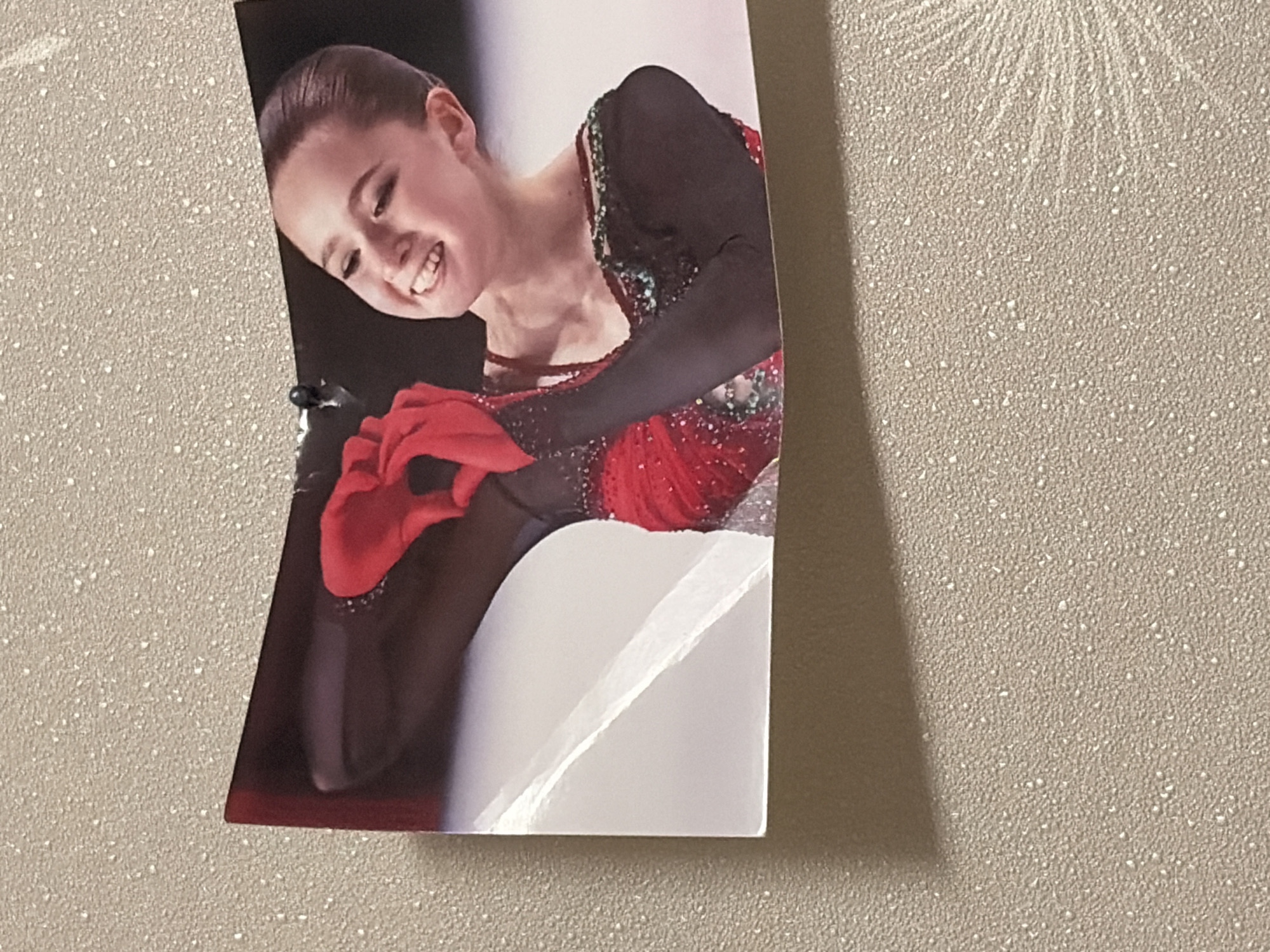 A photograph is displayed on a textured wall, a portion of which is visible in the foreground, showing a grainy, speckled surface in a beige hue, with a shadow cast from above and to the left. The photograph itself is a close-up of a young woman with dark hair pulled back, smiling broadly with her mouth open and teeth visible. She is wearing a dark, long-sleeved garment with shimmering green and red embellishments around the neckline and shoulders, and a red dress with intricate beading over the bodice. Her arms are raised, and her hands, clad in red gloves, are positioned together in front of her chest, forming a heart shape. The background of the photograph appears to be an ice rink with a white surface, suggesting the woman is a figure skater. The photograph is slightly curved, indicating it might be a printed image on flexible material. The location of Leninogorsk, Russia, is not discernible from the visual elements of the image.