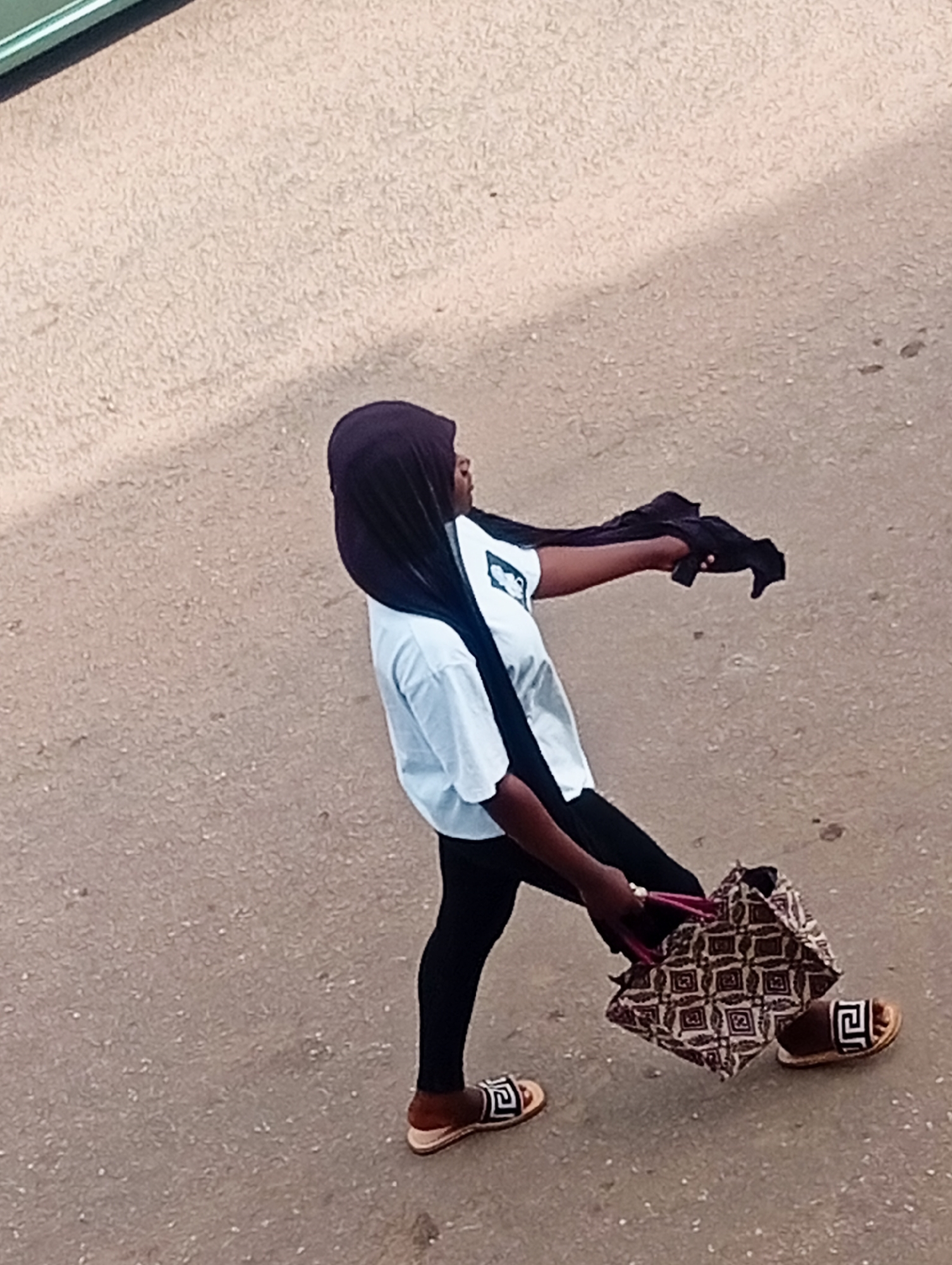 In Bukuru, Nigeria, a person wearing a purple hijab and a light blue t-shirt walks on a paved surface. The person is also wearing black leggings and brown sandals with a black geometric design. In their right hand, they are carrying a patterned bag. Their left arm is extended and holding a dark garment. The ground is a textured, light brown asphalt.