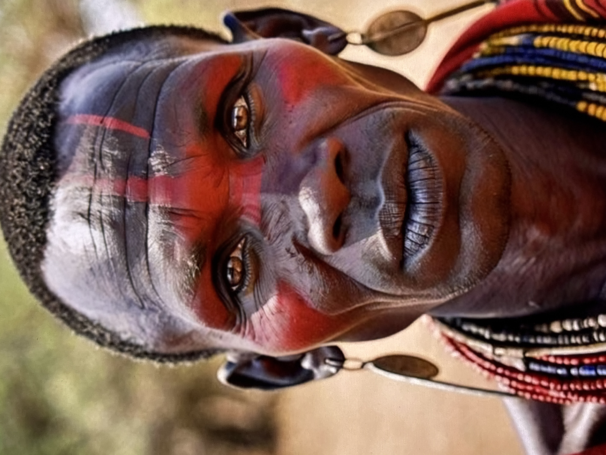 The image is a close-up portrait of a person with dark skin, adorned with red face paint in a striking pattern. The paint features a vertical stripe down the forehead and two horizontal stripes across the cheeks. The person has short, dark hair and is wearing large, oval earrings. Around the neck, multiple strands of colorful beads are visible, predominantly red, yellow, and blue. The background is blurred and appears to be an outdoor environment with earthy tones, suggesting a natural or rural setting. The image does not contain any elements that would indicate a location within the city of Orihuela, Spain.