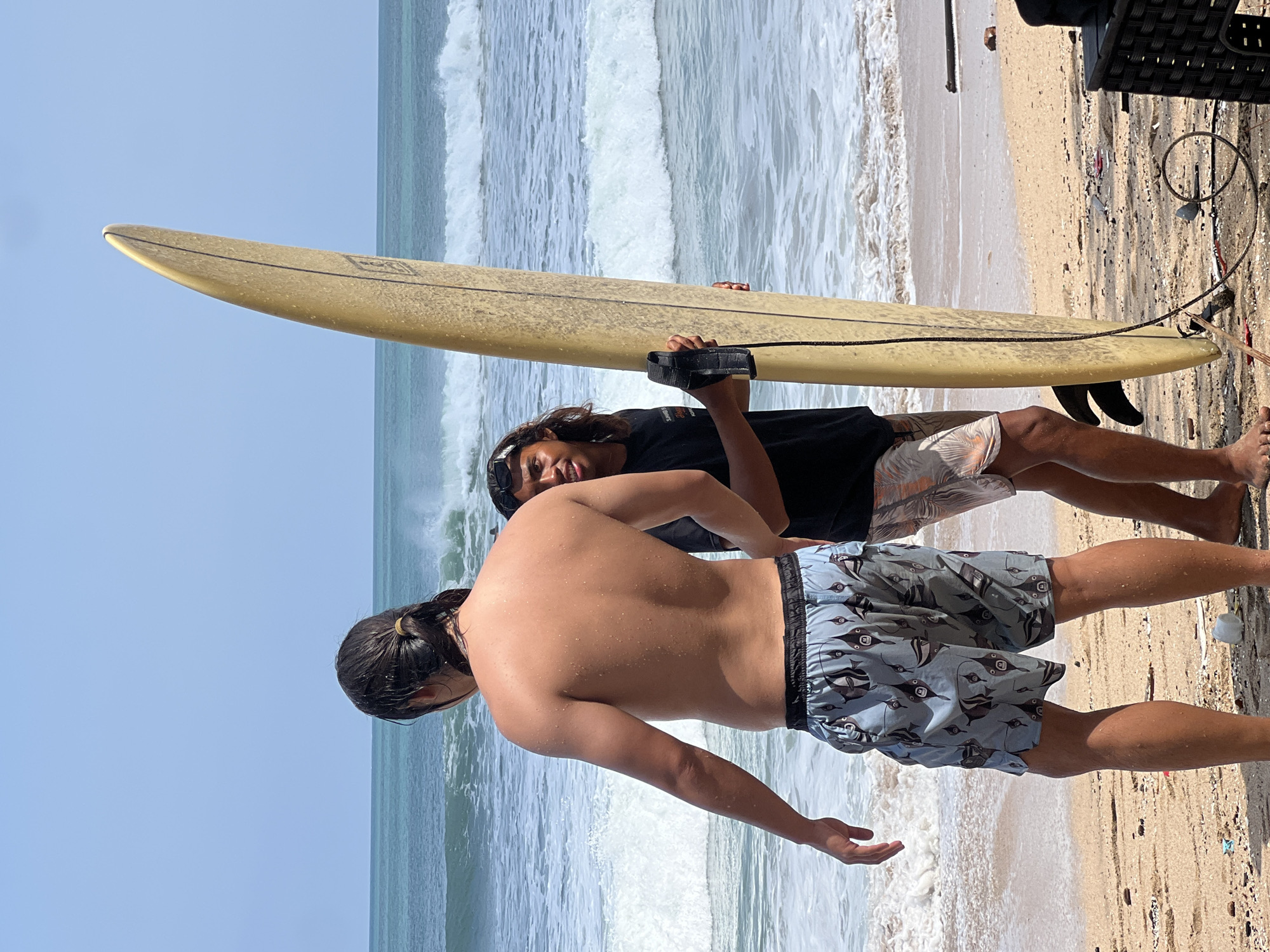 The image depicts a beach scene in Kuta, Indonesia, on a sunny day. Two individuals are present on the sandy shore, facing the ocean. The person on the left, viewed from the back, is a man with dark hair pulled into a ponytail, wearing patterned swim trunks. He appears to be wet. The second individual, on the right, is a man with long, dark hair and sunglasses perched on his head. He is wearing a black t-shirt and patterned swim trunks. He is holding a light-colored surfboard vertically. The ocean in the background shows moderate waves. The sand on the beach is light brown and appears to have some debris scattered on it.