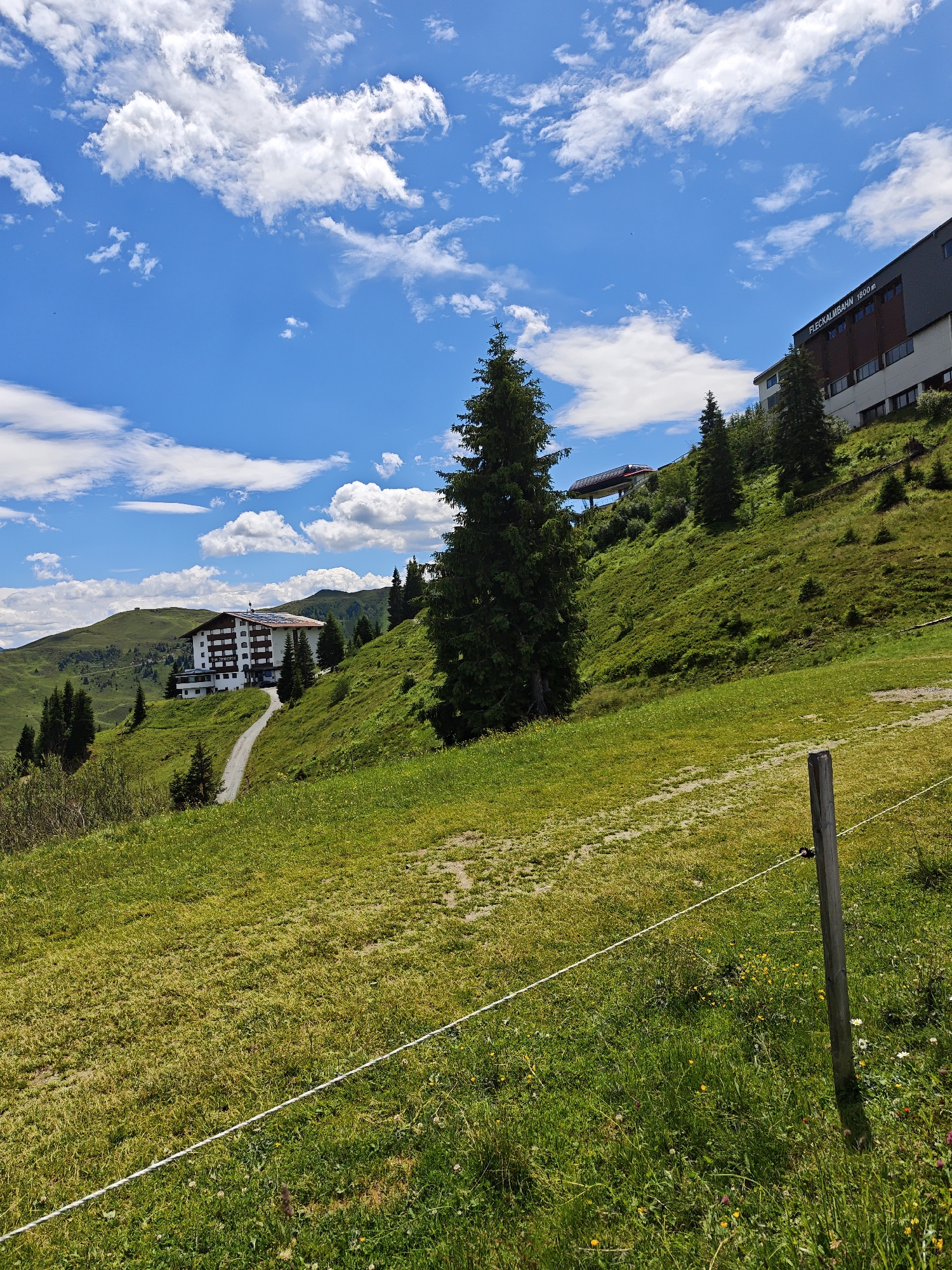 The image depicts a mountainous landscape in Kirchberg in Tirol, Austria, under a partly cloudy blue sky. In the mid-ground, a large, white hotel with a brown roof and solar panels is situated on a grassy hillside. A winding dirt road leads up to the hotel. To the right of the hotel, a tall, dark green evergreen tree stands prominently. Further up the slope, a modern building with a dark facade and the text "FLECKALMBAHN 1800m" is visible, suggesting a cable car station. Another, smaller building with a red roof and a covered structure is also present on the hillside in the upper left quadrant. The foreground consists of a green, grassy field with scattered wildflowers and a white rope fence supported by a wooden post. The overall scene conveys a tranquil, alpine environment.