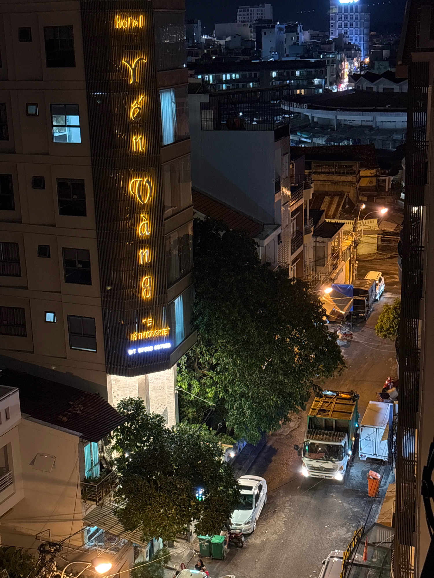 The image depicts a nighttime street scene in Nha Trang, Vietnam. A multi-story building with illuminated yellow neon lettering that spells "HÔTEL YẾN NGỌC" dominates the left side of the frame. The building appears to be a hotel, with windows and balconies visible. Below the hotel's name, additional blue neon lettering reads "TẾ LÝ THƯỜNG KIỆT" and a phone number.
The street below is asphalt and appears somewhat uneven, with some debris or construction materials visible. Several vehicles are present. A white car is parked on the left side of the street. Further down the street, a large garbage truck with its lights on is positioned, facing the camera. To the right of the garbage truck is a white shipping container. An orange trash can is next to the shipping container. A few people are visible on motorcycles and walking.
Adjacent to the hotel, a dense cluster of dark green trees with lush foliage is prominent. In the background, other buildings of varying heights and architectural styles are visible, illuminated by streetlights and internal building lights, creating a typical urban cityscape at night. The overall impression is a busy, functional urban environment, likely a commercial or residential district.