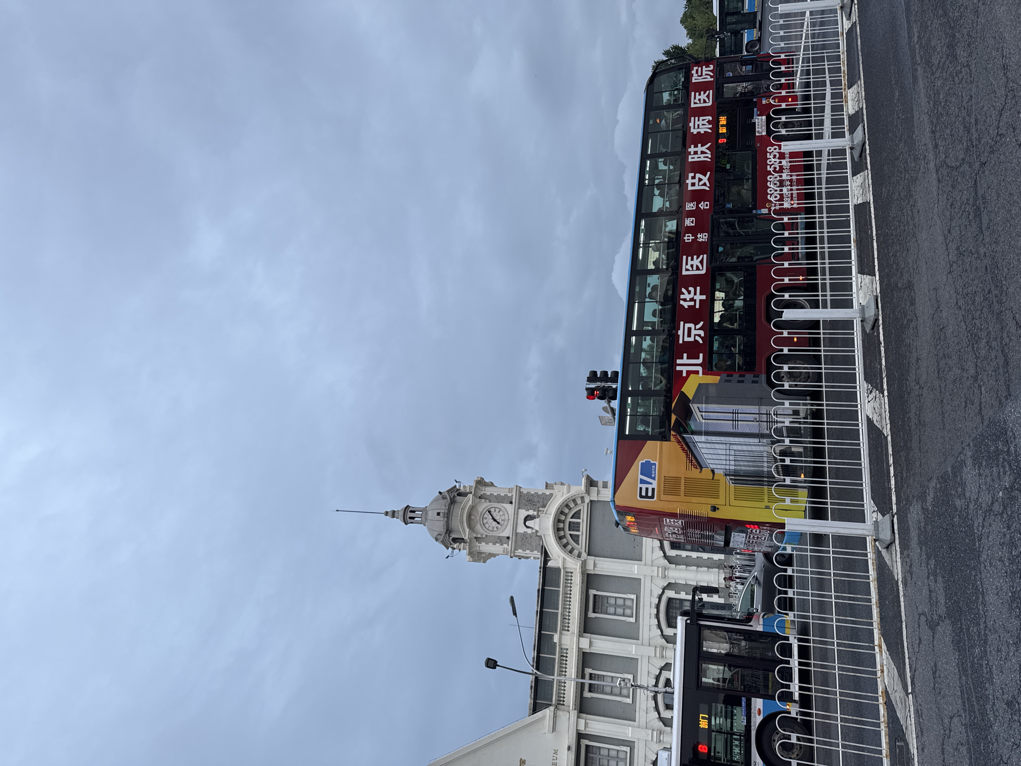 The image shows a street scene in Dongcheng District, China. A clock tower with a spire rises behind a light-colored building. A double-decker tour bus, red and yellow, is parked along the curb in front of a white metal fence. Traffic lights are visible. The sky is overcast. A city bus is also visible.
