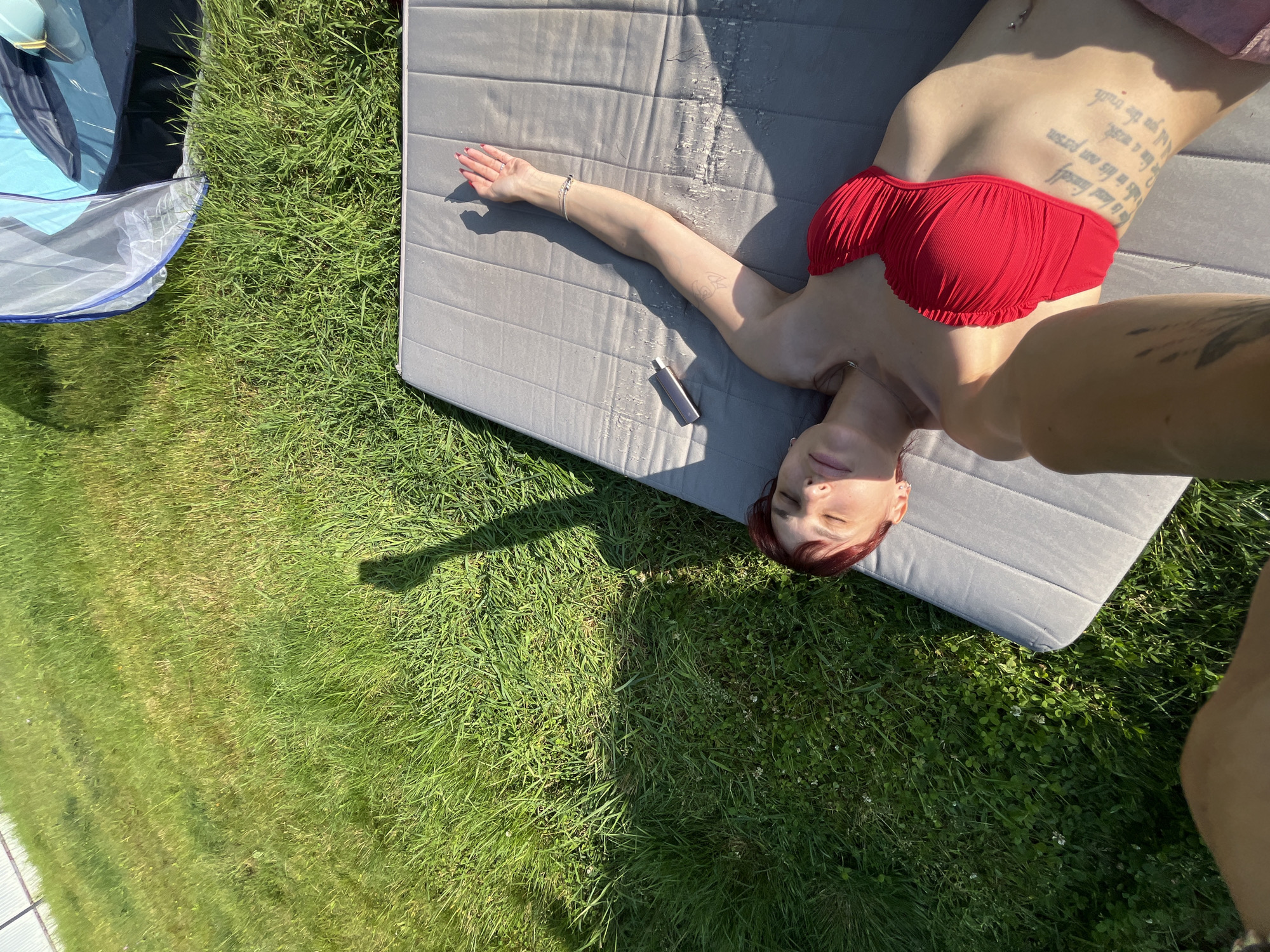 A woman with red hair is lying on a gray mat outdoors. She is wearing a red bikini top and has tattoos on her arm and torso. She is positioned on her back with her head resting on the mat. Her left arm is extended to her side, and her right arm is bent at the elbow, with her hand near her head. A small silver object, possibly a vape or cigarette lighter, is on the mat near her left arm. She is lying on a patch of green grass, and part of a blue and white tent is visible to the left of the frame. The scene appears to be in a park or backyard on a sunny day, with shadows cast by the sun. The location is in the city of Nagorny, Russia.