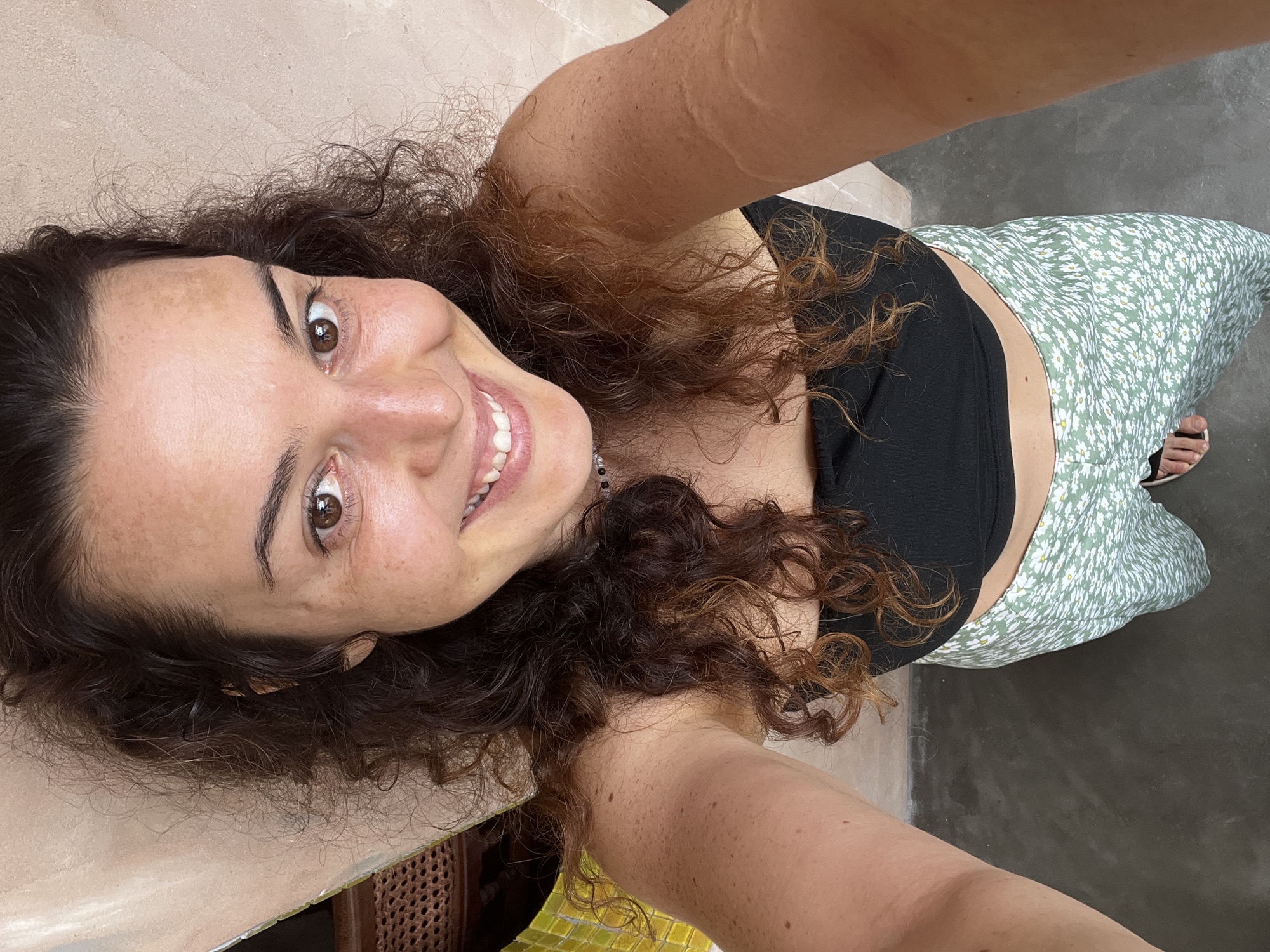 A high-angle, close-up selfie shows a woman with curly brown hair, brown eyes, and fair skin with freckles. She is wearing a black bandeau top and a high-waisted green skirt with a white floral pattern. The woman is smiling, and her mouth is open, revealing white teeth. The background consists of a textured, light-colored wall on the right and a dark, concrete floor on the left. The scene appears to be outdoors, possibly in a courtyard or patio area, given the concrete floor and the wall texture. The woman is in the foreground, occupying the majority of the frame. The overall impression is a casual, candid shot. There are no identifiable landmarks or specific details that would definitively place this scene in Mũi Né, Vietnam.