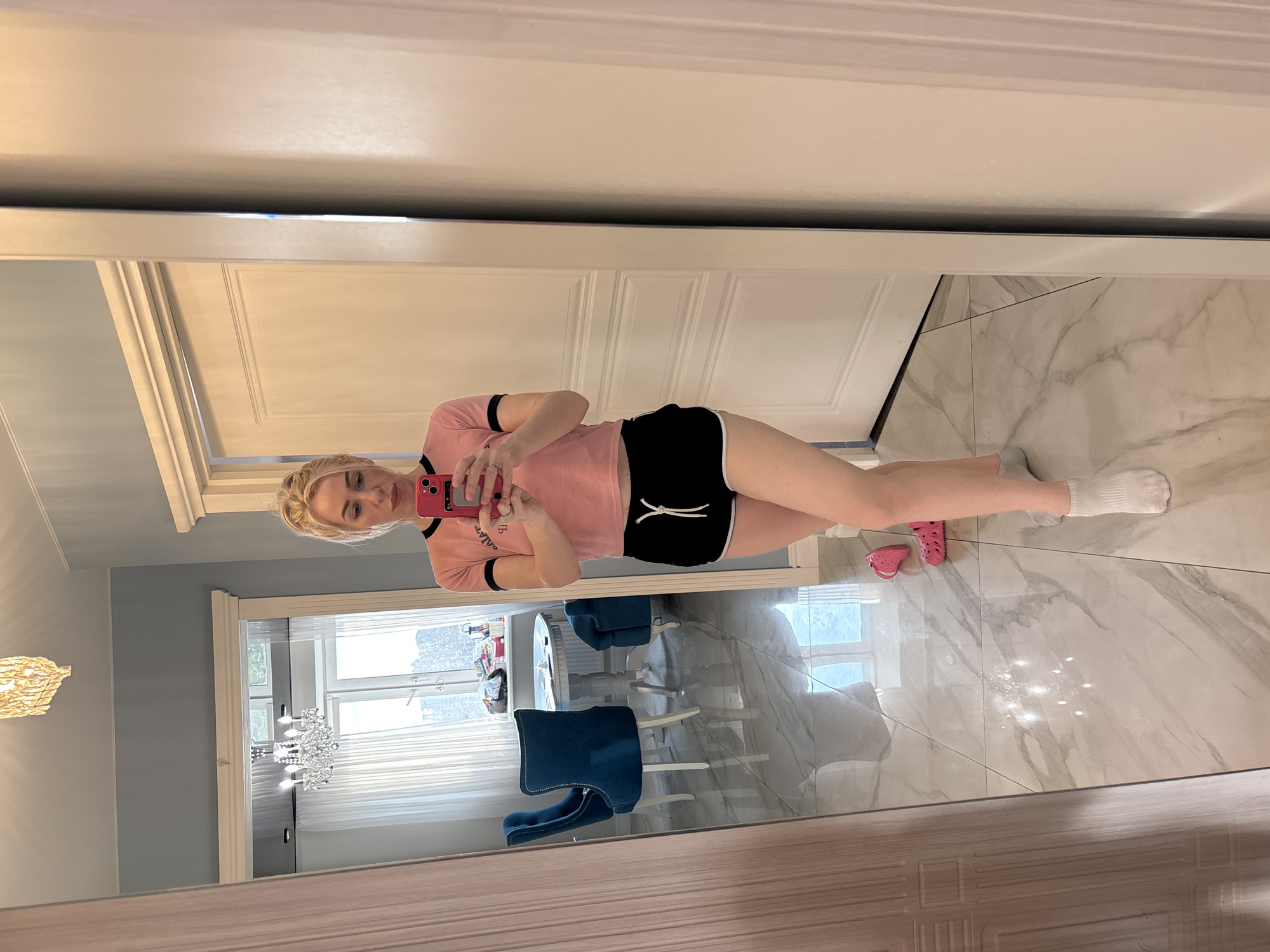 A woman is taking a selfie in a mirror. She is wearing a pink t-shirt with black trim, black shorts with white ties, and white socks. There are pink Crocs on the floor near her feet. The floor is white marble. In the background, a dining room is visible with a chandelier, white walls, blue chairs, and a table set with dishes. The scene is located inside a home, potentially in Sinyavino, Russia.