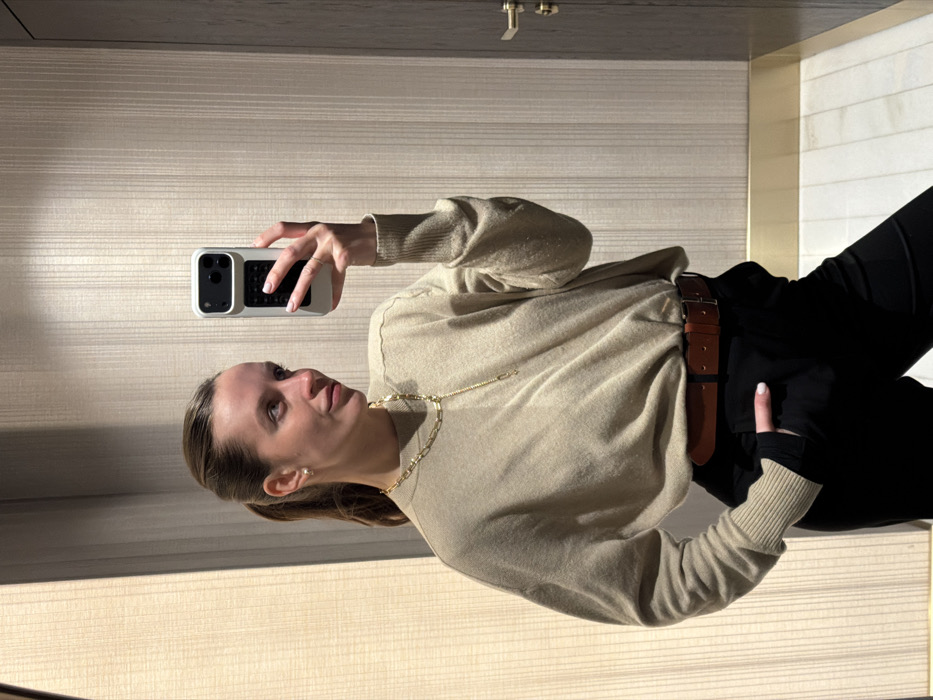 A female individual is depicted indoors, taking a mirror selfie with a light-colored smartphone in the city of Budva, Montenegro. The subject, an adult female with light brown hair pulled back into a ponytail, wears a beige long-sleeved sweater, black trousers, and a brown leather belt. She is accessorized with a gold-colored chain necklace with a Y-drop design and small stud earrings. Her left hand holds the smartphone, which has a textured dark grip on its case, while her right hand rests in her trouser pocket. She faces towards her right with a slight smile. The background features a light-colored wall with vertical paneling, and a dark wooden door with a brass handle is partially visible on the right. The floor is composed of light-colored tiles, separated from the wall by a gold-colored trim. The scene is evenly lit by ambient indoor lighting.