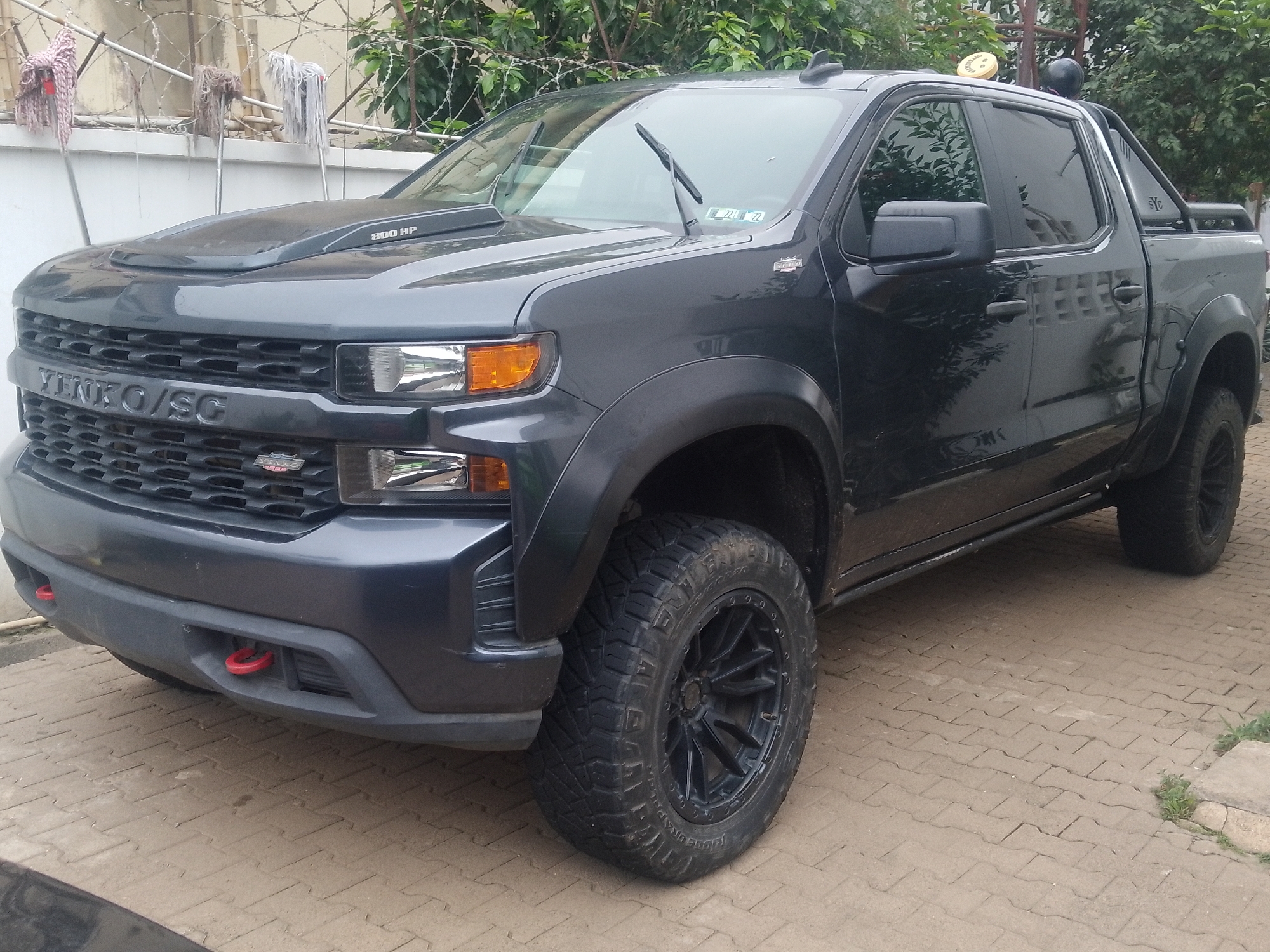A dark grey Chevrolet pickup truck, modified with off-road tires and wheels, is parked on a paved walkway in Abuja, Nigeria. The truck features a distinctive grille with "YENKO/SC" branding, a hood scoop, and red tow hooks on the front bumper. In the background, a white wall with barbed wire fencing is visible, along with lush green trees and some mop heads hanging on poles. The truck is positioned at an angle, showing its front and the driver's side. The overall scene suggests a residential or semi-urban environment within Abuja.