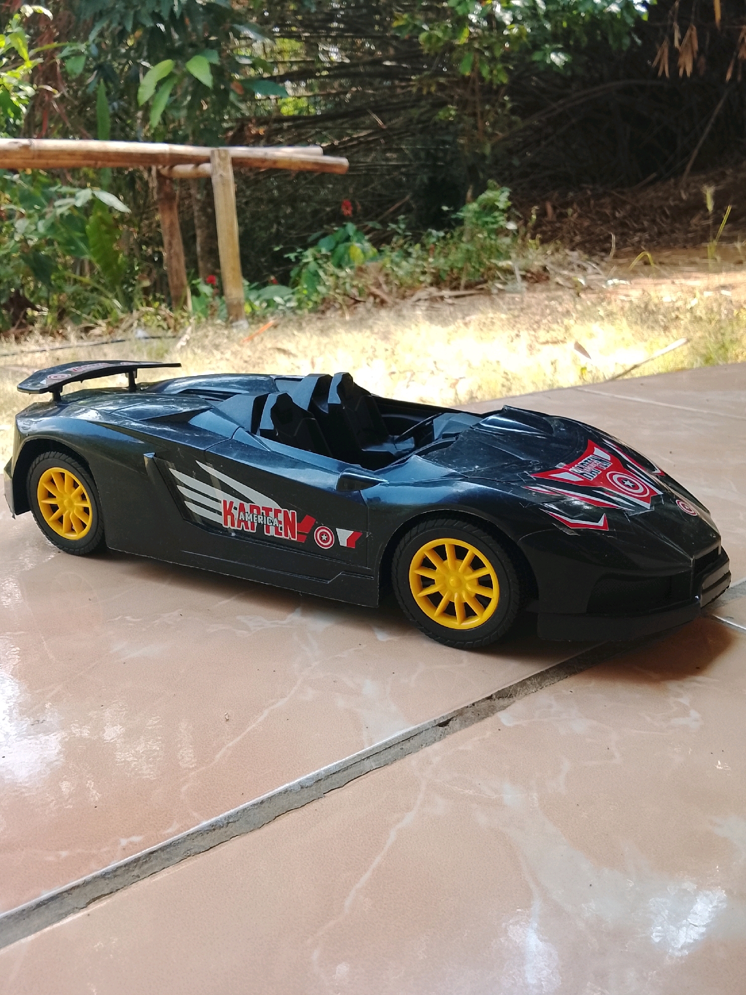 The image shows a black toy sports car with yellow wheels positioned on a tiled surface. The car has decals on its sides, including the word "KARTEN" and a circular emblem that resembles the Captain America shield. The background of the image is blurred, but appears to be an outdoor setting with greenery and bamboo structures. The lighting suggests it is daytime. This scene is located in Kertahayu, Indonesia.