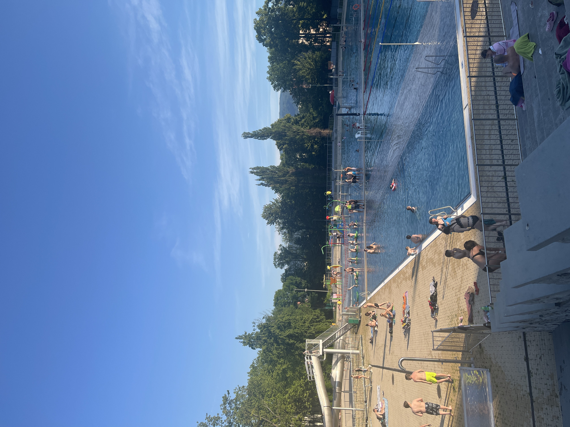 An outdoor swimming pool facility is shown on a clear day with a bright blue sky. The pool area is filled with numerous people of various ages, many of whom are swimming, playing in the water, or relaxing on the poolside. There are different sections of the pool, including a large main pool with lane markers, a shallower area with play structures, and a slide. The surrounding area features paved walkways, loungers, and grassy areas. Trees and buildings are visible in the background, suggesting an urban or suburban setting. The lighting and shadows indicate it is late afternoon or early evening.