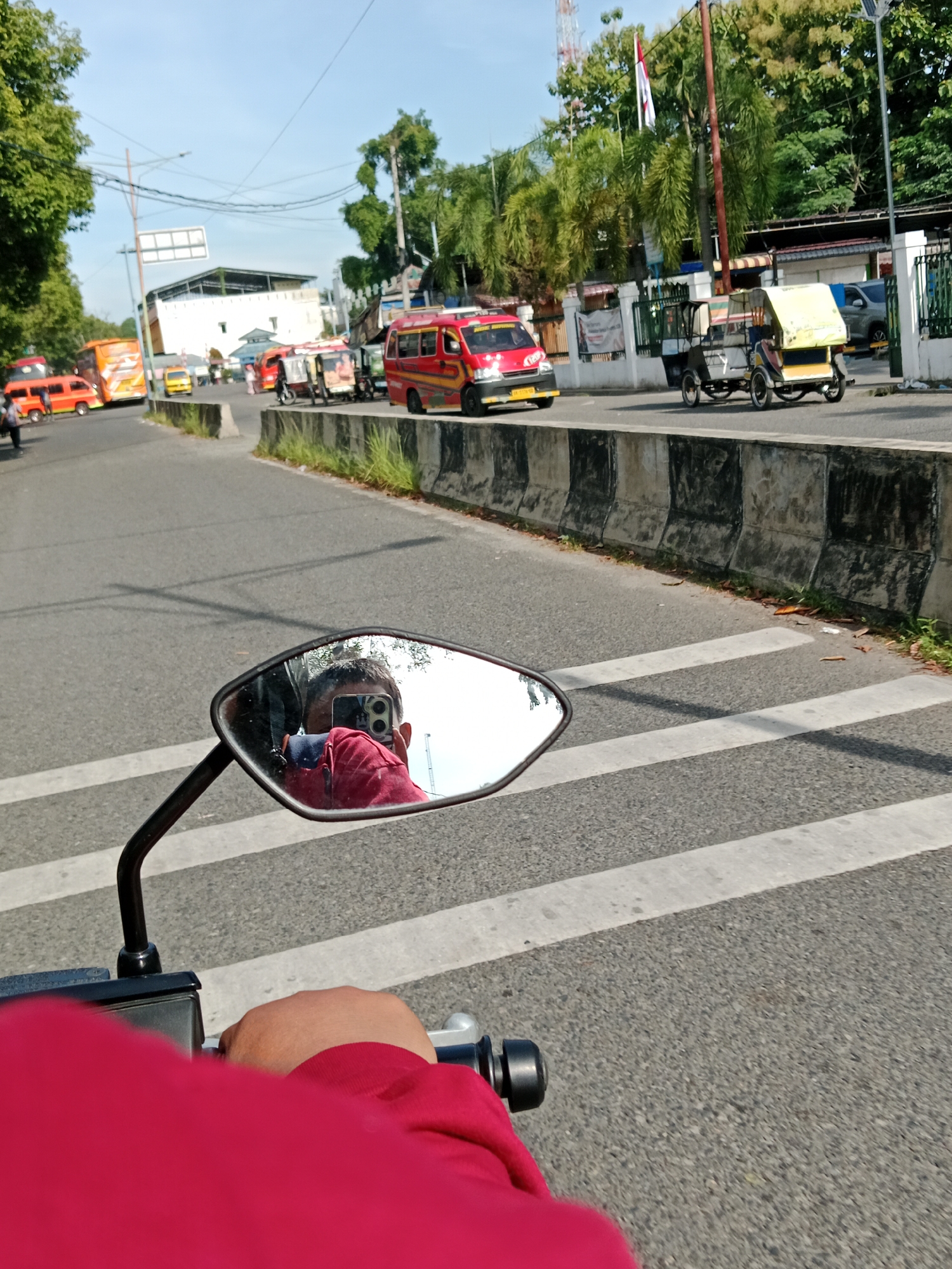 The image captures a street scene in the City of Binjai, Indonesia, likely during daylight hours as indicated by the bright sky and shadows. The perspective is from a person riding a motorcycle, with their hand on the handlebar visible in the foreground and a portion of their red shirt. A side-view mirror attached to the motorcycle reflects the rider's face and upper body, who is holding a smartphone to take a selfie. The rider appears to be wearing a red shirt and possibly a helmet, though it's obscured by the reflection.
The street is paved and marked with white pedestrian crossing lines. Several vehicles are present on the road, including a red minivan, a yellow minivan, and what appears to be a trishaw or becak with a yellow canopy. A larger orange bus is visible further down the road. The environment includes a concrete barrier along the side of the road, some vegetation, trees, and buildings in the background, including a tower with a flag. The overall scene depicts a typical urban street with traffic and pedestrians.