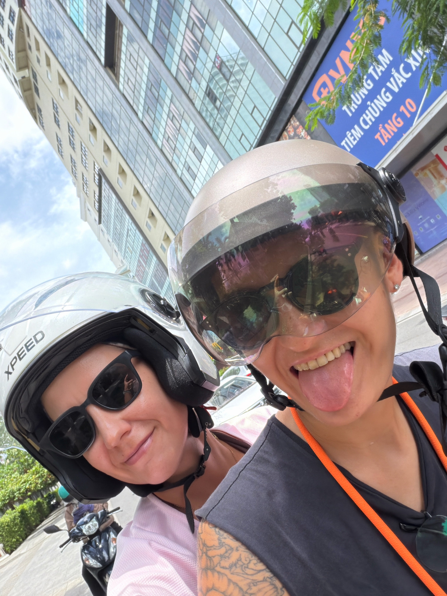Two individuals, one in the foreground and one slightly behind, are riding a scooter in Nha Trang, Vietnam. The person in the foreground wears a light-colored helmet with a reflective visor and sunglasses, sticking their tongue out playfully. They are wearing a dark t-shirt with an orange cord around their neck. The person behind them wears a white helmet with "XPEED" visible and dark sunglasses, smiling at the camera. They are wearing a pink top. The background features a modern building with many windows and a blue sign with Vietnamese text, likely indicating a business or establishment. Trees and other foliage are also visible, suggesting an urban environment. The overall scene captures a moment of casual enjoyment during a scooter ride.