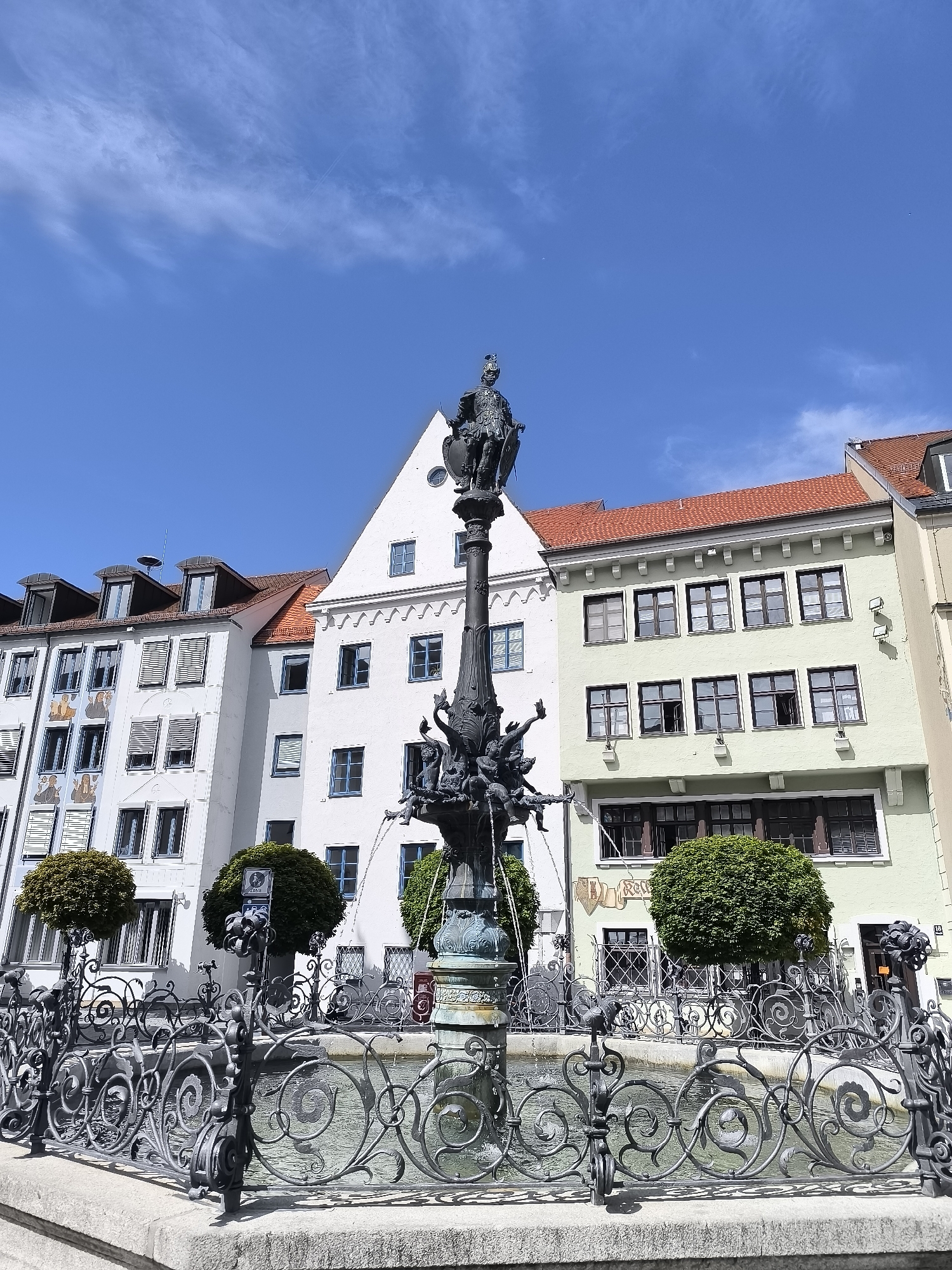 In the city of Kempten (Allgäu), Germany, a black metal ornate fountain stands in a public square. The fountain features a central column with a statue of a knight at its peak, surrounded by several smaller figures. Water sprays from the fountain into a shallow basin. The basin is enclosed by an intricately designed black metal fence with swirling patterns and decorative elements. Behind the fountain, a row of buildings with traditional architecture lines the square. The buildings have varying facade colors, including white, light green, and grey, with red tiled roofs. Some buildings feature multiple windows with blue shutters or frames, and one building displays artwork on its facade. Green, manicured bushes are planted around the fountain area. The sky above is a clear, vibrant blue with wispy white clouds and a faint airplane trail. The scene captures a bright, sunny day in a European city.