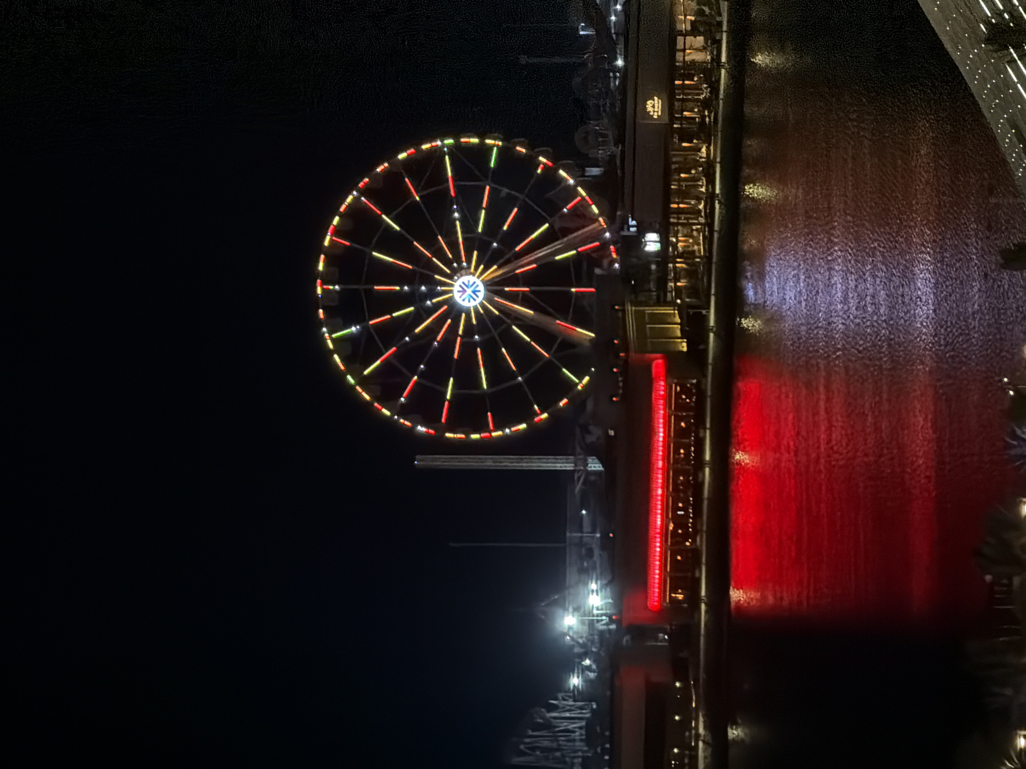 A Ferris wheel illuminated with colorful lights stands tall against a dark night sky, its spokes extending outwards from a central hub. Below the Ferris wheel, a waterfront promenade is lined with buildings featuring illuminated signage and windows. The water in the foreground reflects the vibrant red lights from the buildings, creating a shimmering effect. In the background, the silhouettes of roller coaster tracks and other amusement park structures can be seen. The scene suggests an entertainment district or amusement park.