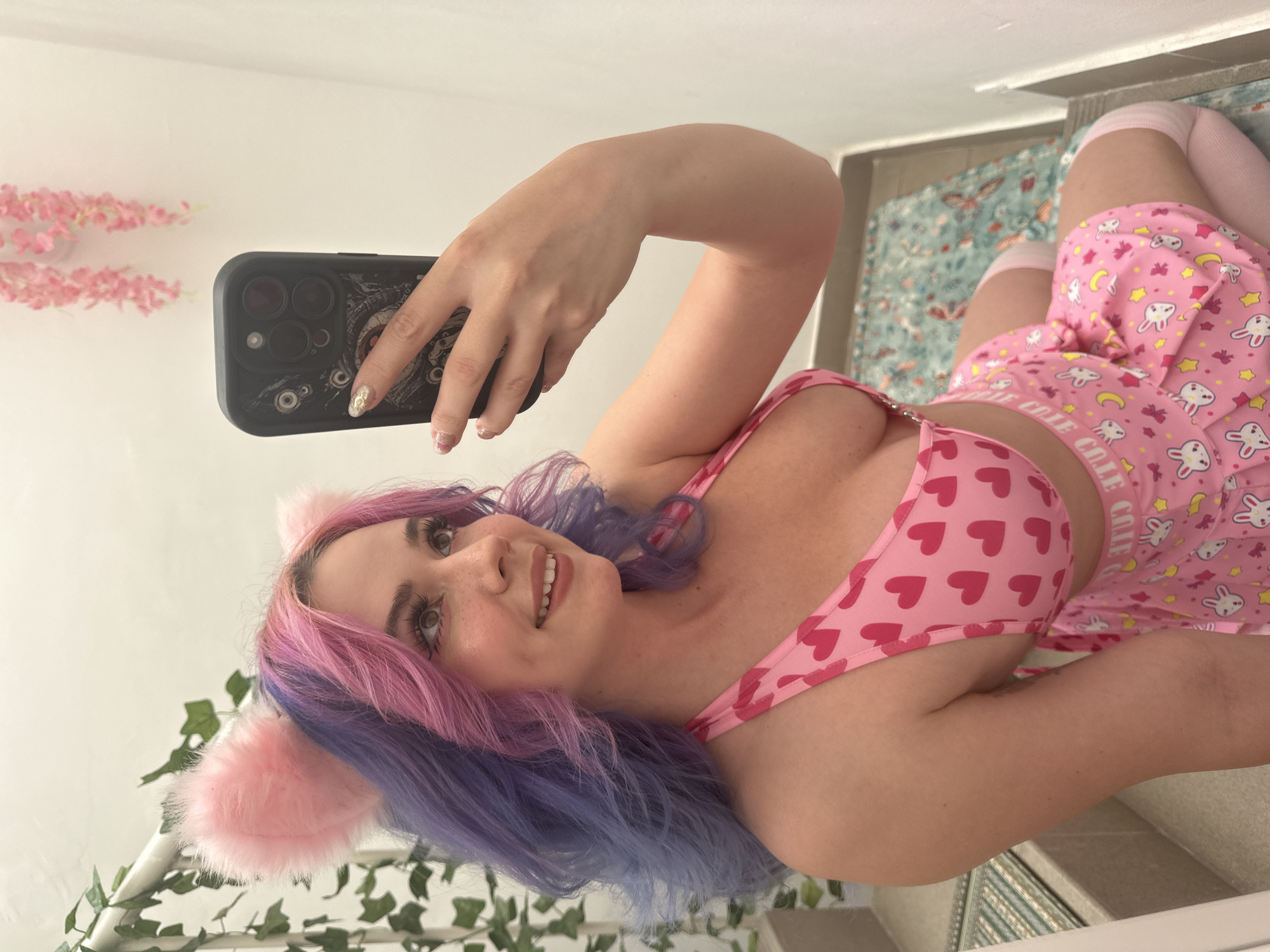 A person with pink and purple hair is wearing pink cat ears and a matching pink outfit with hearts and bunny patterns. The person is holding a phone with a dark case in their hand, taking a selfie. There is a flowering vine and a leafy vine visible in the background. A colorful rug can also be seen in the background. The location is likely indoors and not in the City of Zagreb, Croatia.