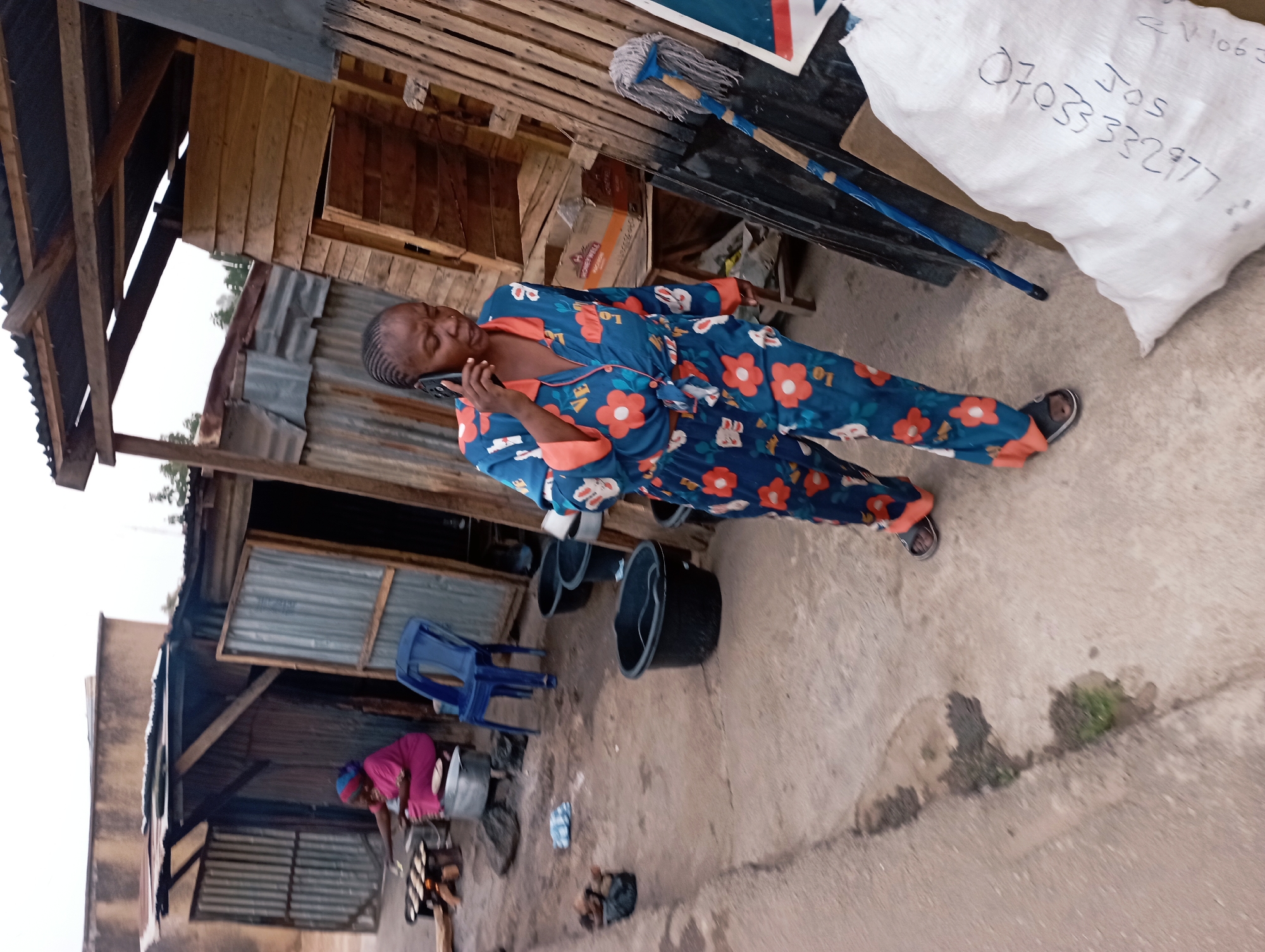 An African woman in floral pajamas and slippers is walking and talking on a cell phone in an outdoor setting. Behind her are makeshift structures with wood and corrugated metal walls and roofs. A seated woman in a purple shirt is tending to a small stove. Black buckets are on the ground. A large white bag with writing that includes "JOS" and a phone number "07033332977" is visible near the woman on the phone. The scene appears to be in a residential area of Jos, Nigeria.