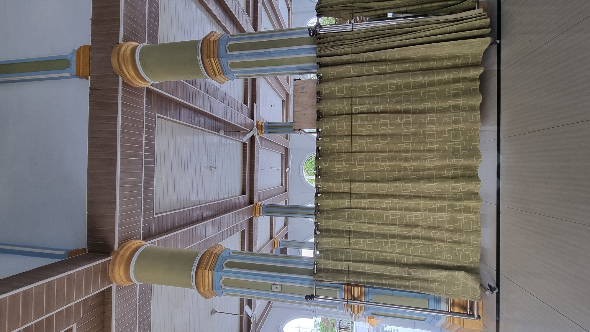 This is an interior photograph taken from a low angle, looking upwards at a colonnade and ceiling structure. Four large, fluted columns, painted in alternating bands of green, blue, and gold, support a wooden plank ceiling with white trim. The ceiling is divided into rectangular sections with a repeating pattern of wooden planks and white paneling. Two ceiling fans are visible in the distance. A long, olive-green curtain with a repeating geometric pattern of white squares hangs across the lower portion of the image, obscuring the view of what lies behind it. The floor is tiled with large, light-colored rectangular tiles. The photograph appears to have been taken in Meunasahlhok, Indonesia, suggesting a public or religious building, possibly a mosque or community hall, given the architectural style. There are no people or discernible actions in the frame.