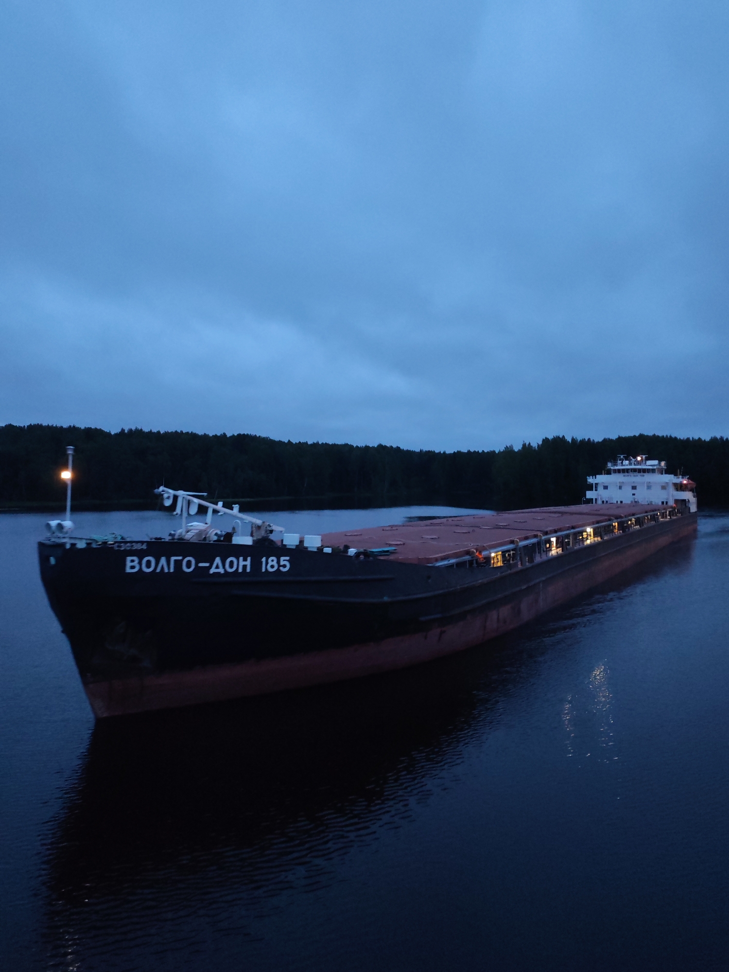 An aerial view captures a large cargo barge, identified by the Cyrillic text "ВОЛГО-ДОН 185" emblazoned on its hull, navigating a dark, calm body of water. The barge is seen from a slightly elevated perspective, revealing its elongated deck covered in what appears to be red tarpaulin. The vessel's superstructure, a multi-story white building, is positioned at the stern, illuminated by interior lights. Numerous smaller lights are visible along the sides of the cargo deck.
The surrounding environment is dominated by a dense, dark forest that forms a continuous line along the distant shoreline, contrasting with the dimly lit sky, which shows signs of cloud cover. The water reflects the barge's lights and hull, creating a subtle shimmering effect. The overall atmosphere is one of dusk or early morning, with low light conditions. There are no visible individuals on the deck. The image is devoid of any urban elements, suggesting the location is remote and not within the city of Markovo, Russia, but rather in a natural waterway environment.