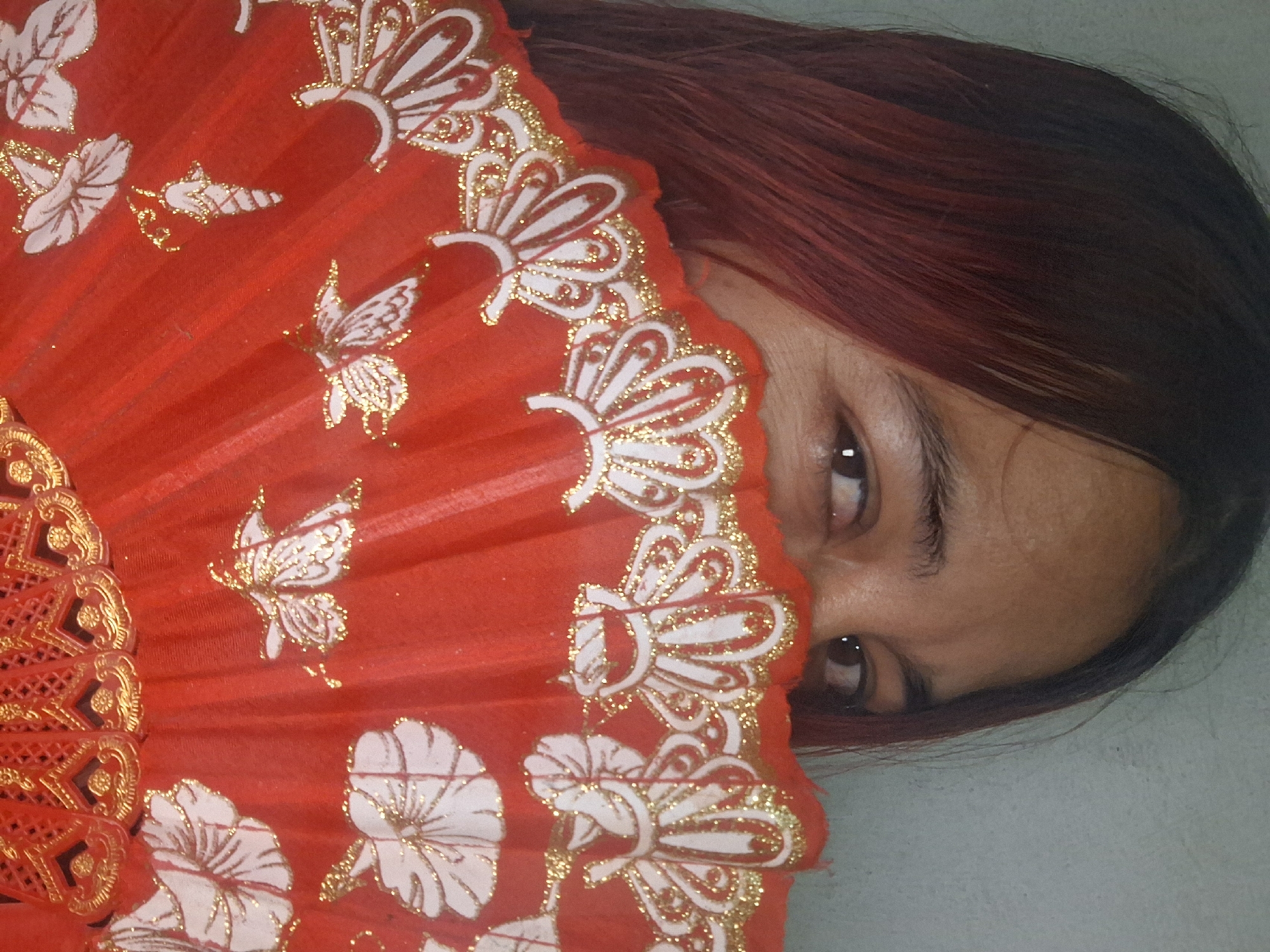 A person with dark hair dyed reddish-brown is partially obscured by a red folding fan with white and gold detailing. The fan features floral and butterfly designs. The person's eyes are visible above the fan. The background is a plain, light gray surface. The location is unknown, but the city is specified as Vallecito, Venezuela.