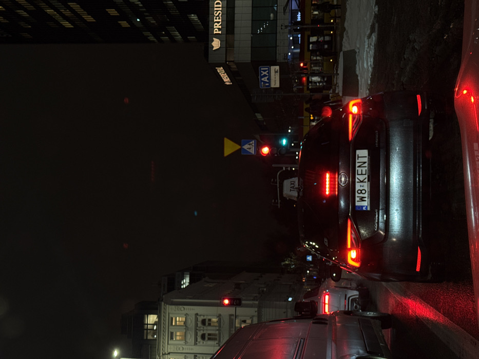 A nighttime scene depicts a busy intersection in Warsaw, Poland, specifically in the vicinity of the West Station Office Center at Aleje Jerozolimskie 123A. The viewpoint is from inside a vehicle, indicated by a red reflection in the lower foreground.
The environment is dark, illuminated by streetlights and vehicle lights. The road surface is wet, reflecting red light, while patches of snow are present on the right-hand curb and sidewalk.
In the immediate foreground, a dark-colored Toyota Corolla Hybrid sedan, registered with Polish license plate "W8 KENT," is stopped. It displays illuminated red taillights and a roof-mounted "TAXI" sign. Directly to its left, a white van and a white SUV are also stopped, their red taillights visible.
Multiple traffic signals are illuminated, displaying red for vehicular traffic. Above the central traffic signals, a yellow inverted triangle "yield" sign and a blue square "pedestrian crossing" sign are present.
On the left side of the street, a multi-story building with classical architectural elements features several illuminated windows. On the right, a modern skyscraper with numerous illuminated windows rises above a lower commercial structure. This lower building prominently displays "PRESIDENTIAL" with a crown logo, identifying it as the West Station Office Center. Another "TAXI" sign is affixed to this building.
Further right, a yellow and red tram is visible, and several indistinct figures, presumed to be pedestrians or passengers, are present on the adjacent sidewalk and tram platform.