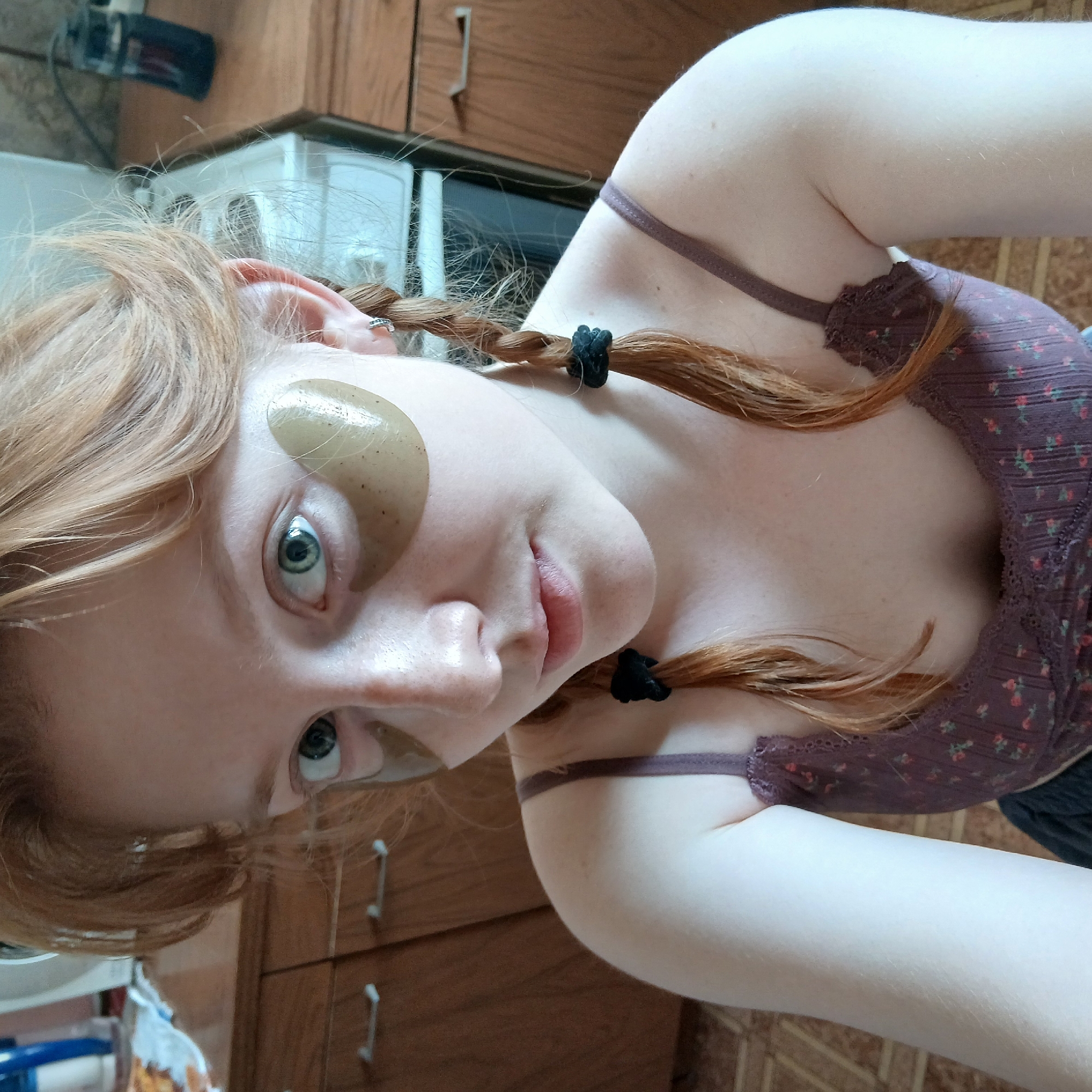 An indoor portrait depicts a fair-skinned young person, likely in Yekaterinburg, Russia, engaged in a skincare routine. The subject has long, reddish-blonde hair styled in two pigtails or braids, secured with black elastic ties. Hydrogel eye patches are applied beneath both of their light-colored, wide-open eyes, which are directed towards the camera. A small, simple earring or cuff is visible on the upper helix of their left ear.
The person is wearing a dark purple or maroon camisole, possibly a bralette, featuring a subtle floral pattern and delicate lace trim along the neckline. Their skin shows a scattering of freckles, particularly on the chest and arm. The overall expression is neutral, with a hint of serenity or curiosity.
The background suggests a home environment, possibly a kitchen, with warm-toned wooden cabinets or drawers forming a dominant feature. Parts of a white appliance, resembling a refrigerator or microwave, are also visible. The lighting is soft and ambient, characteristic of an indoor daytime setting. The image is taken at an oblique angle, with the subject's head tilted, creating a dynamic composition. There is no visible text in the image.