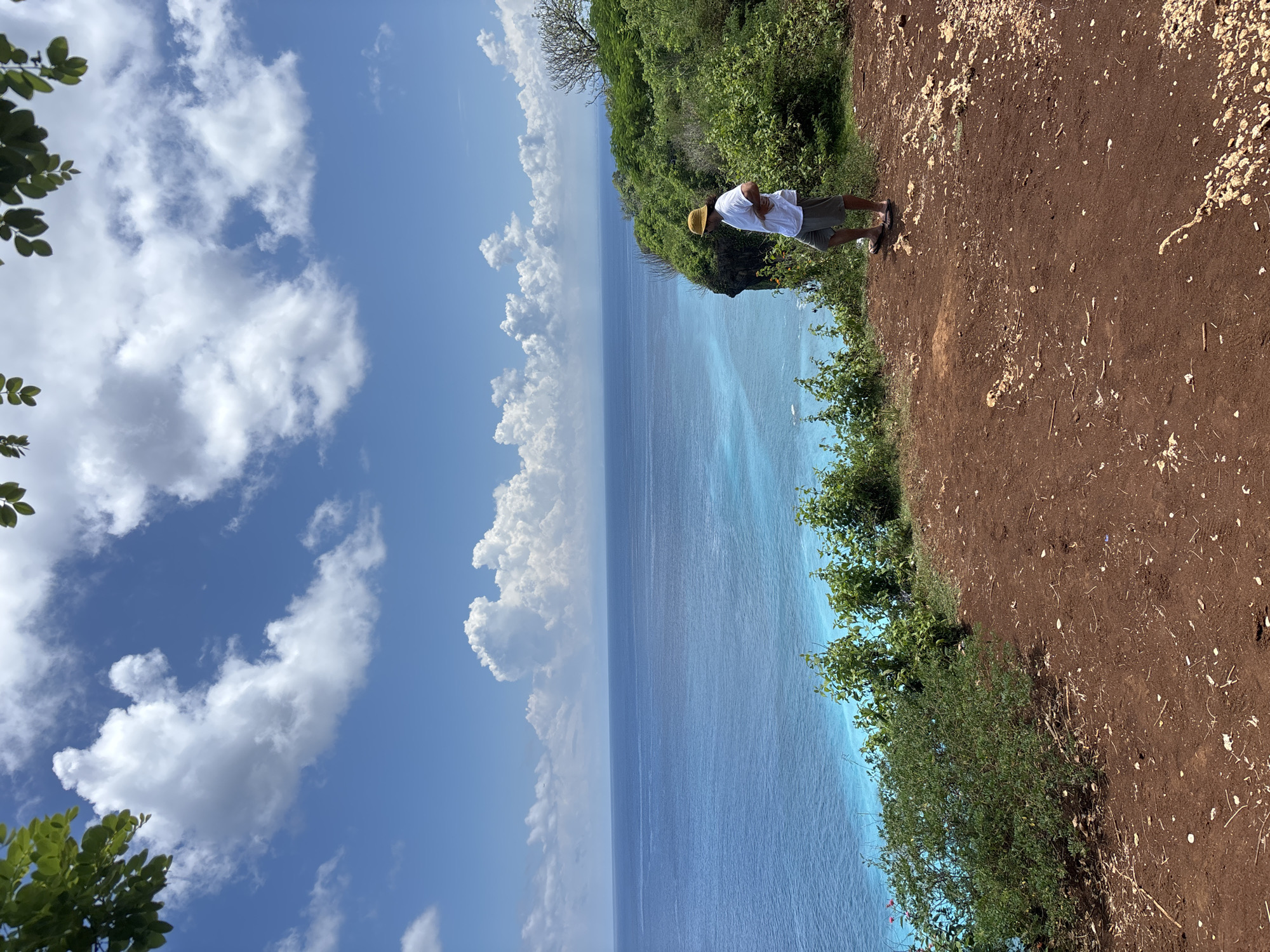 A vertical landscape shot shows a man standing on a high, brown dirt cliff overlooking a blue ocean and sky filled with puffy white clouds. The man is wearing a white short-sleeved shirt, khaki shorts, and a straw hat. He stands near the edge of the cliff, looking down at the ocean. Green vegetation grows along the cliff edge. The blue ocean stretches to the horizon, meeting a blue sky filled with clouds. Uluwatu, Indonesia is likely the location given the cliffside views of the ocean.