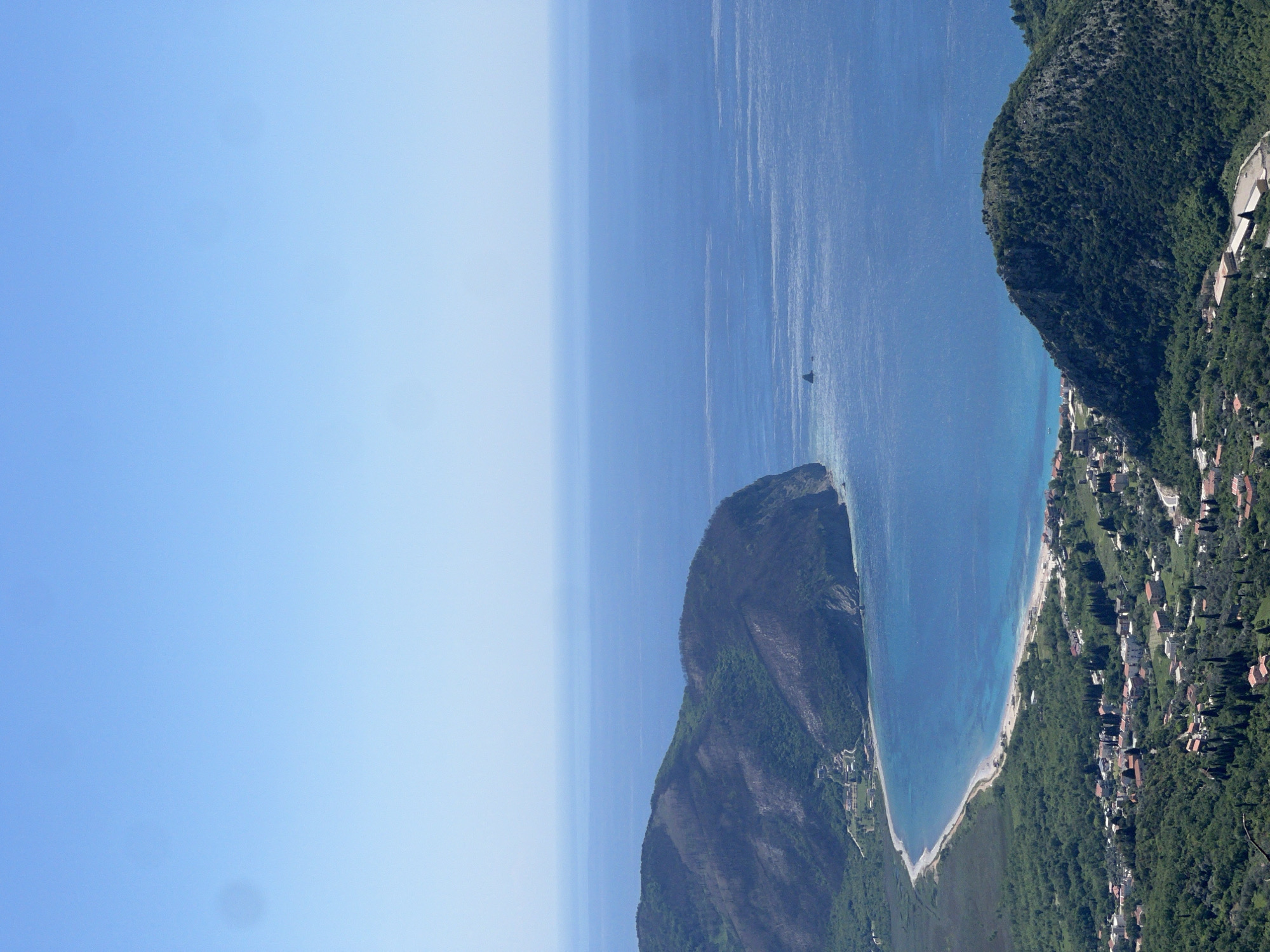 This high-angle, panoramic photograph captures a serene coastal landscape near Novoselje, Montenegro, on a clear, sunny day. The scene is dominated by a vast expanse of bright blue sky at the top, transitioning smoothly into a lighter shade towards the horizon. Below, a wide stretch of deep blue sea shimmers with subtle reflections of sunlight, indicating calm waters.
A prominent feature is the rugged, green-forested coastline that frames the sea. To the right, a large, densely wooded mountain or hill slopes down, meeting a long, curving, light-colored beach that stretches along the water's edge. Along the lower slopes and at the base of this mountain, a scattered settlement is visible, comprising numerous houses and buildings with red or light-colored roofs nestled amidst the verdant trees. Another forested headland or peninsula juts into the sea further along the coast.
A small, dark-colored boat can be seen sailing in the open water, appearing as a solitary detail on the expansive sea. The overall impression is one of natural beauty and tranquility, with the vibrant blue of the sky and sea contrasting beautifully with the rich green of the land. The lighting suggests it is late morning or early afternoon, with no clouds in the sky. There is no visible text in the image.