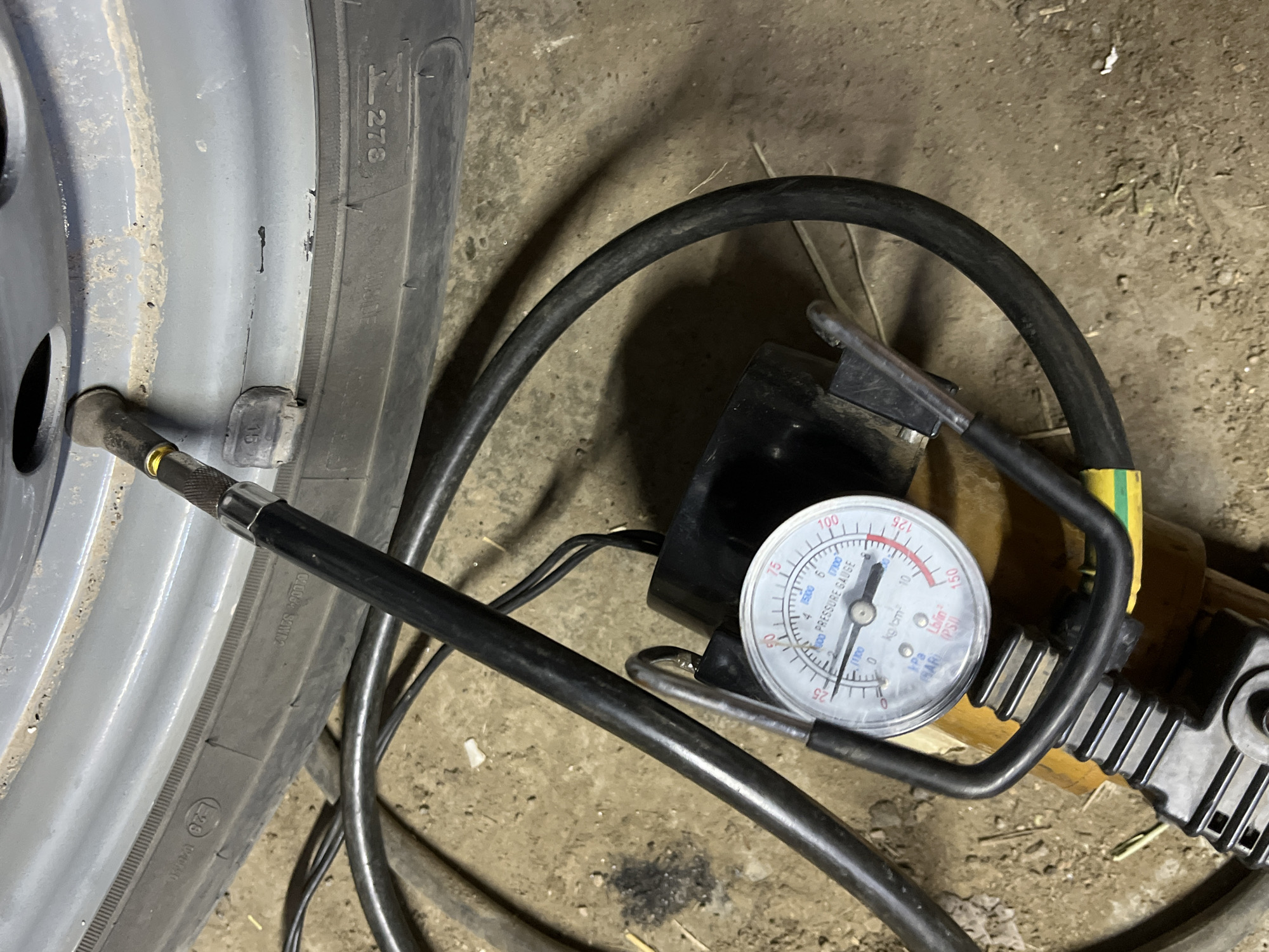 In the city of Krasnyye Barrikady, Russia, a close-up shot captures a vehicle's tire being inflated. The tire's sidewall displays the markings "275" and some illegible text. A black air hose, connected to an air compressor with a yellow and black casing, is attached to the tire's valve stem. A pressure gauge, with a white face and black markings, is integrated into the hose assembly, showing a reading between 0 and 25 psi. The gauge also indicates pressure in kgf/cm², kPa, and lb/in². The scene is set on a dirt or gravel floor, suggesting an outdoor or garage environment.