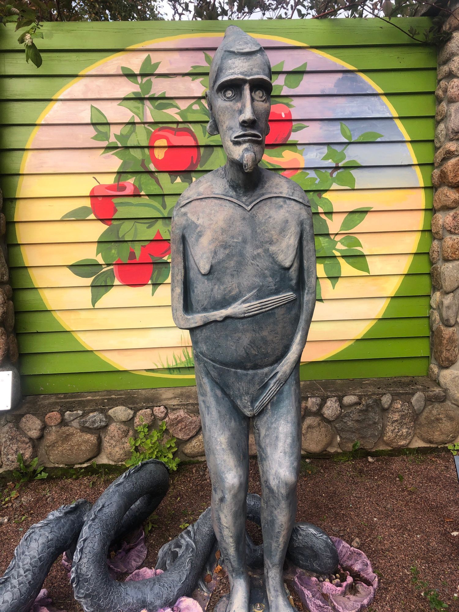 In the city of Inturist SNT, Russia, a statue of a stylized human figure stands prominently in front of a green wooden wall adorned with a mural of apples on a branch within a yellow circle. The statue is made of a dark, weathered material, possibly metal or stone, and depicts a person with a stern expression, wearing a pointed head covering and what appears to be a loincloth. The figure's hands are positioned across its abdomen. A segmented, serpentine form, also dark and textured, is coiled around the statue's legs and base. The ground surrounding the statue is covered in reddish-brown gravel, with scattered green plants and rough-hewn stones forming a border behind the statue and along the muraled wall. The overall impression is of an outdoor art installation with a mythological or symbolic theme.